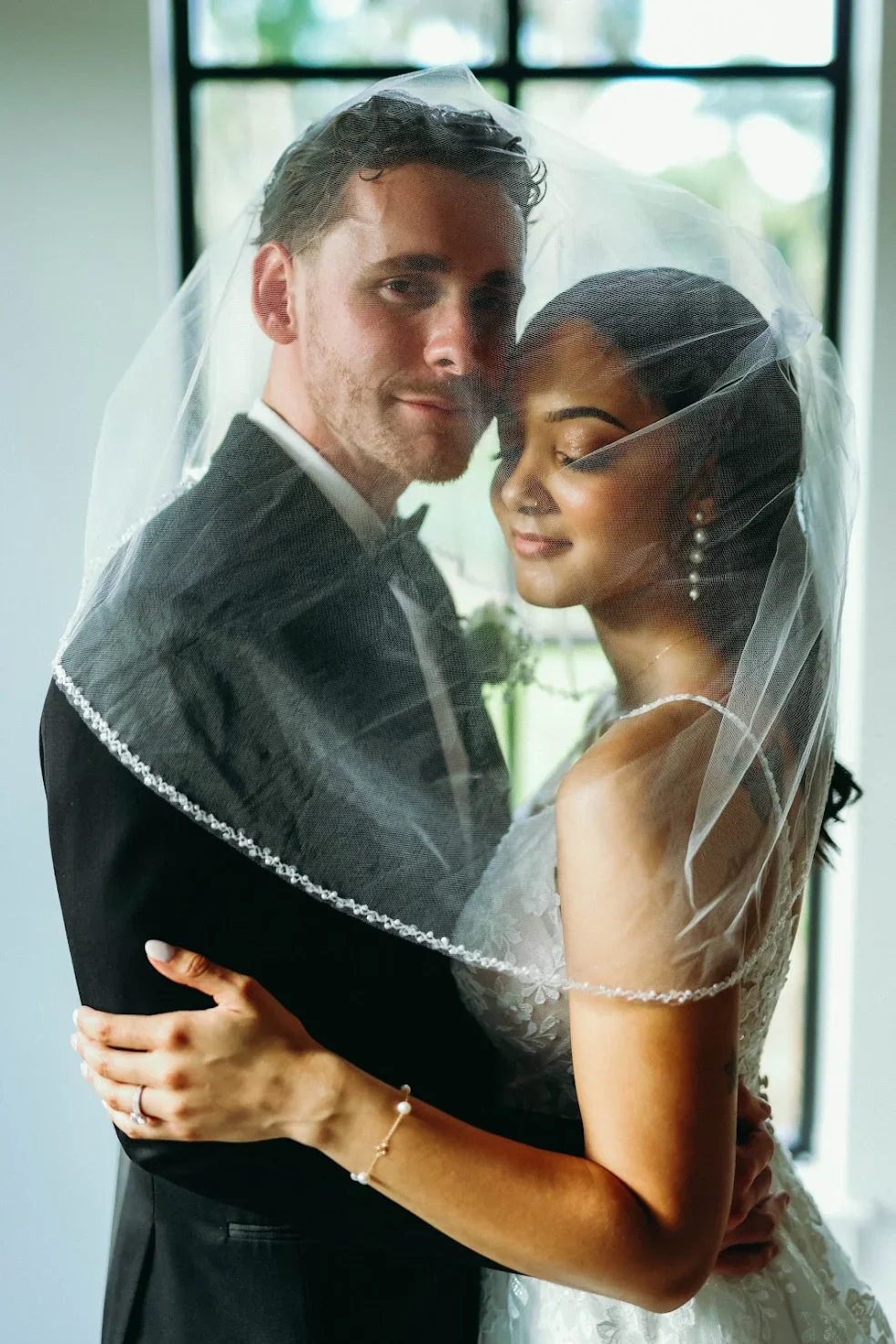 A newlywed couple embracing, with the bride wearing a white wedding gown and veil, and the groom in a black tuxedo, standing near a window with natural light.