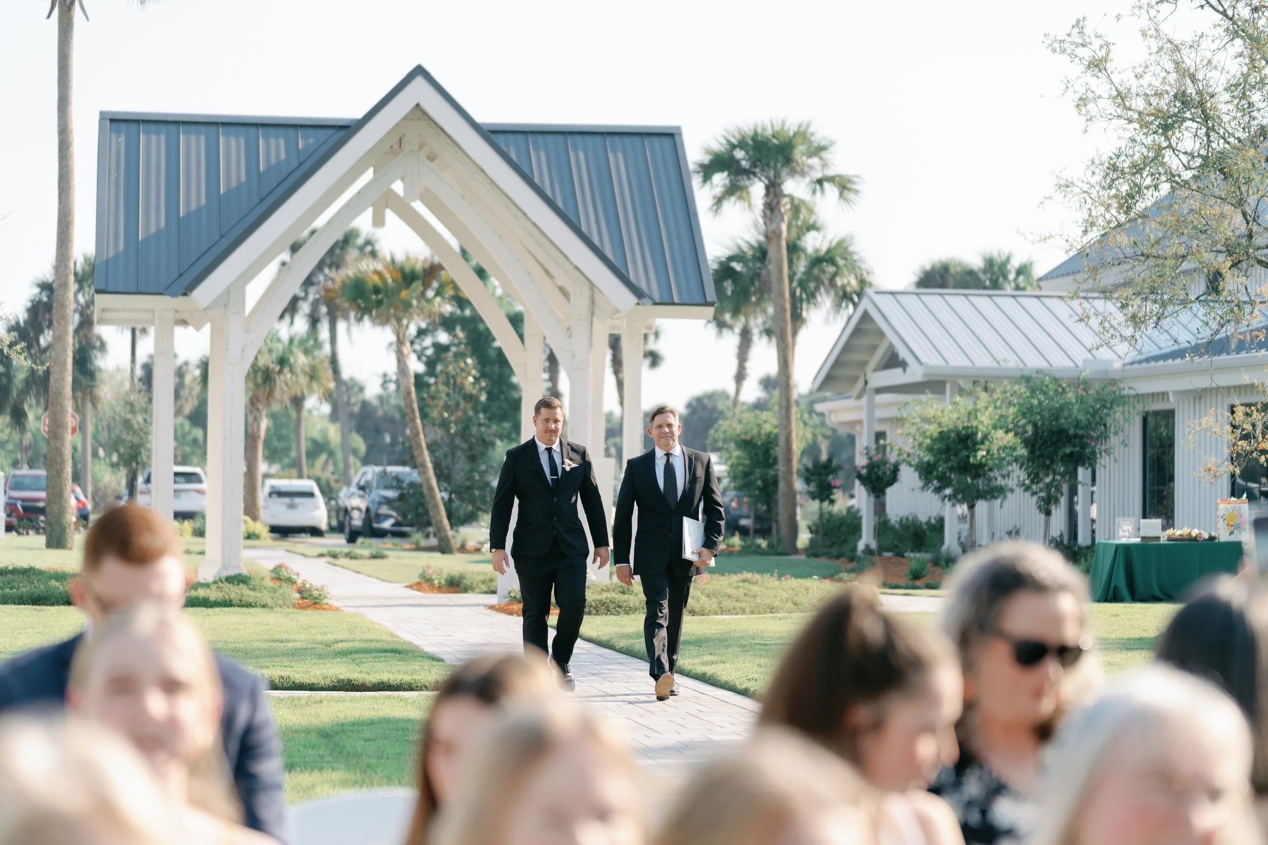Two men in black suits walking on a brick path at an outdoor event, with guests in the foreground and white building with palm trees in the background.