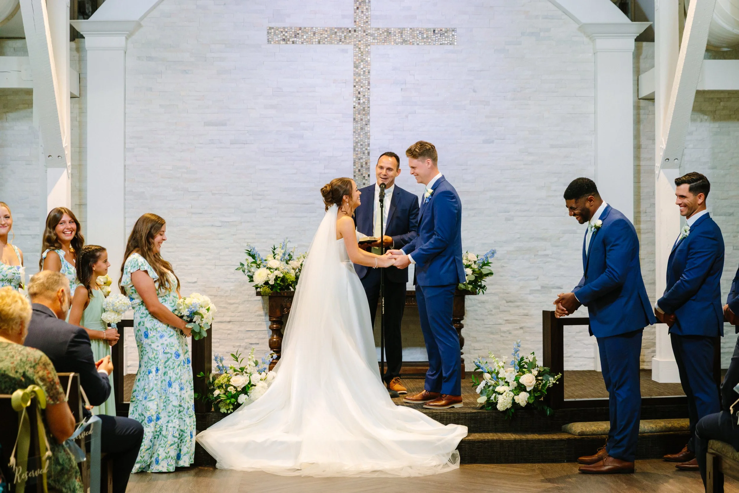 A wedding ceremony inside a church with a large cross on the white brick wall. The bride in a white gown and the groom in a blue suit are holding hands, exchanging vows, with an officiant standing between them. Bridesmaids and groomsmen stand on eith