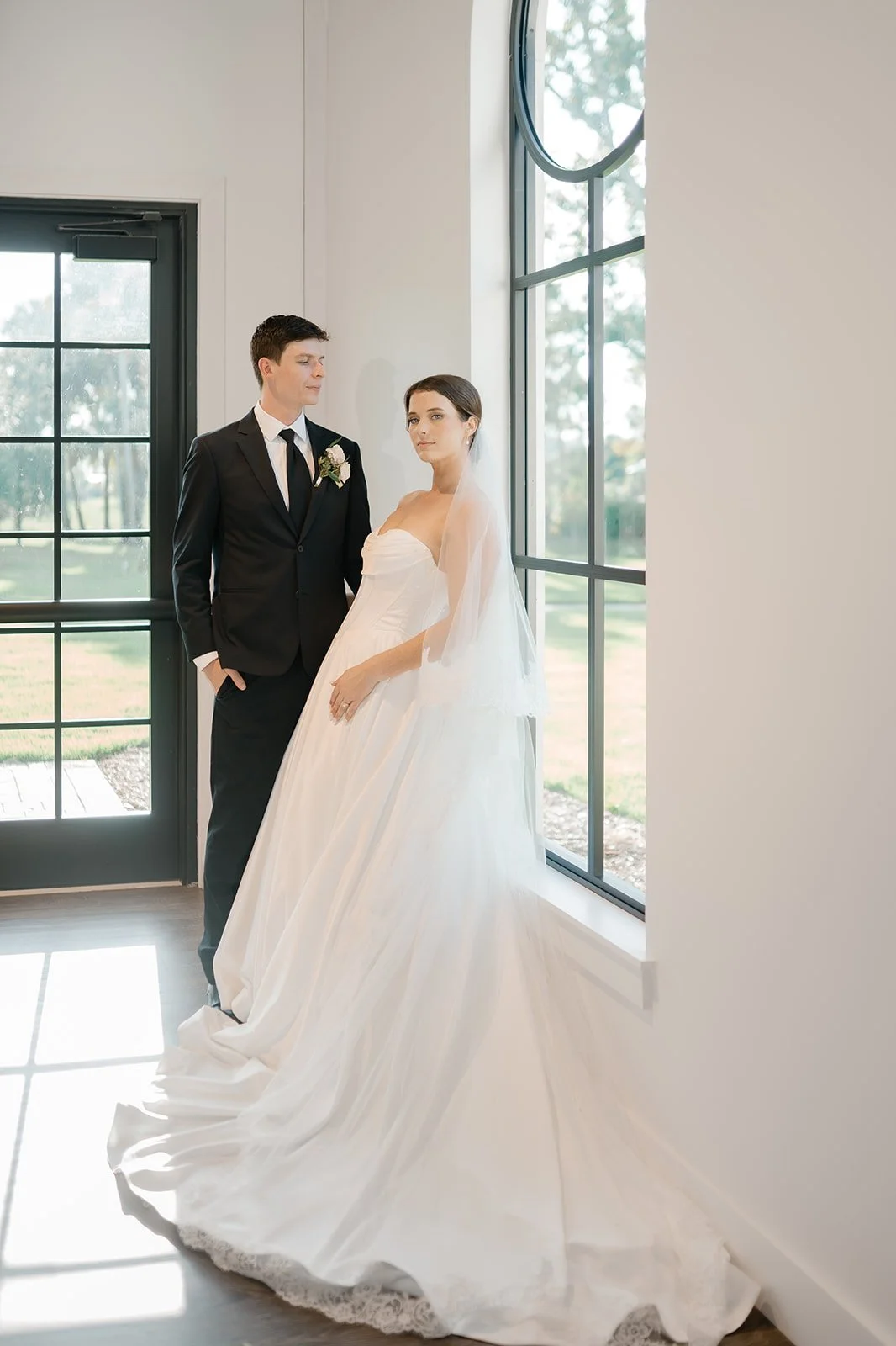 A bride and groom standing indoors near a large window during their wedding, with the bride in a white wedding dress and veil, and the groom in a black tuxedo.