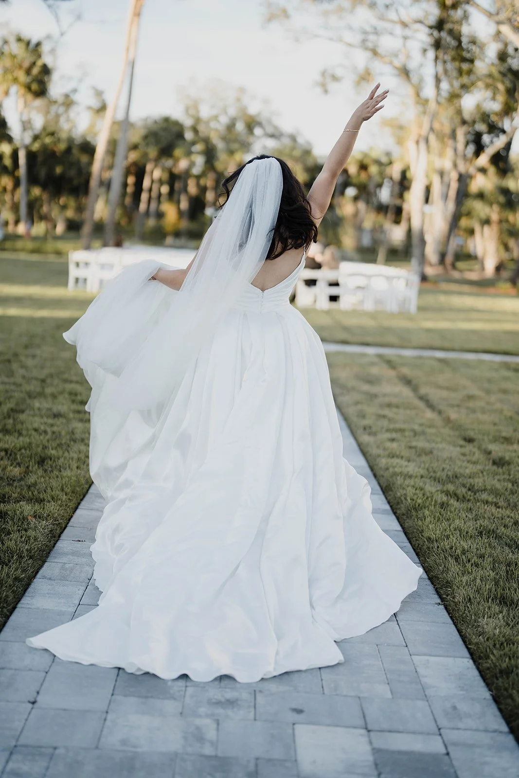 Bride in a white wedding dress and veil dancing outdoors on a stone pathway, with trees and chairs in the background.