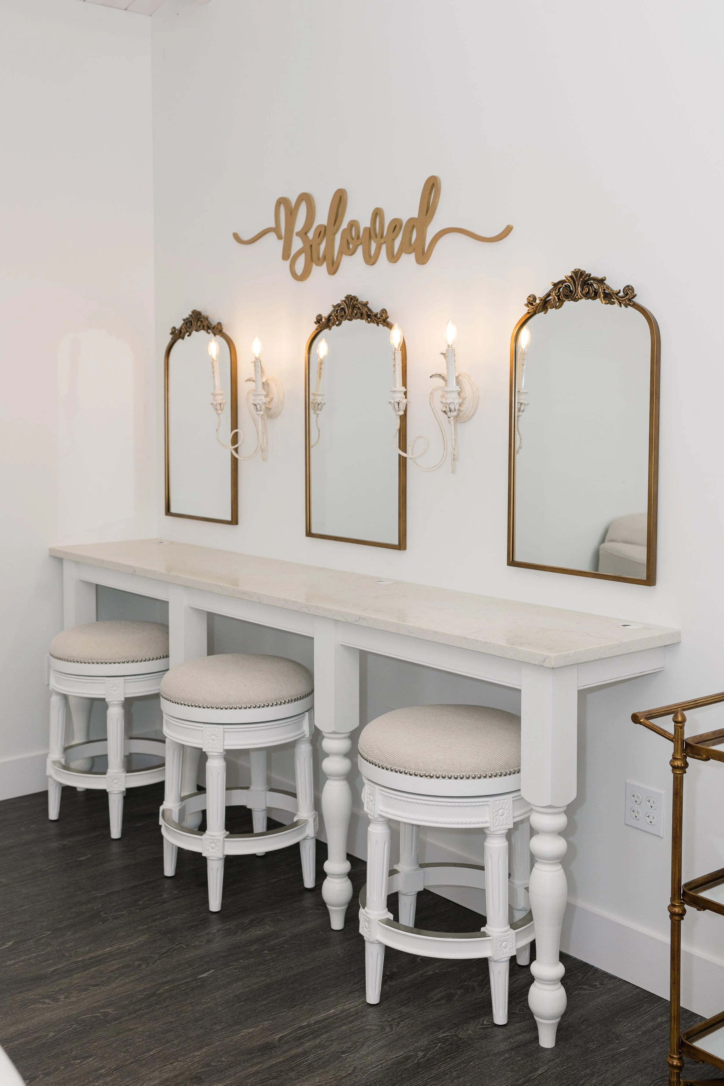 White vanity with three upholstered stools, three mirrors with ornate gold frames, wall sconces with candles, and a gold sign that says 'Blessed'