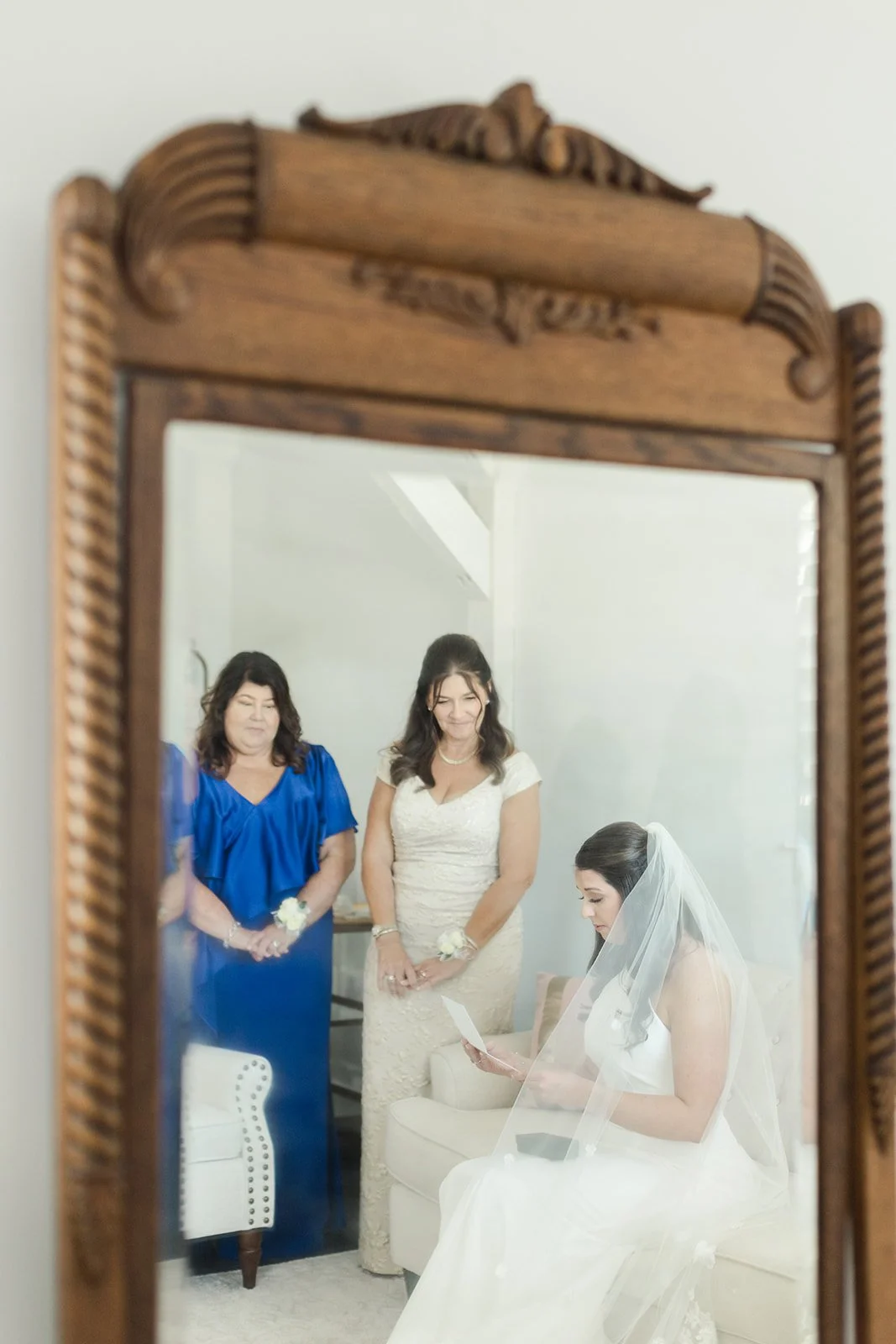A bride in a white wedding dress with a veil sitting on a couch and reading a letter, reflected in a mirror. Two women in formal dresses stand behind her, looking at her.