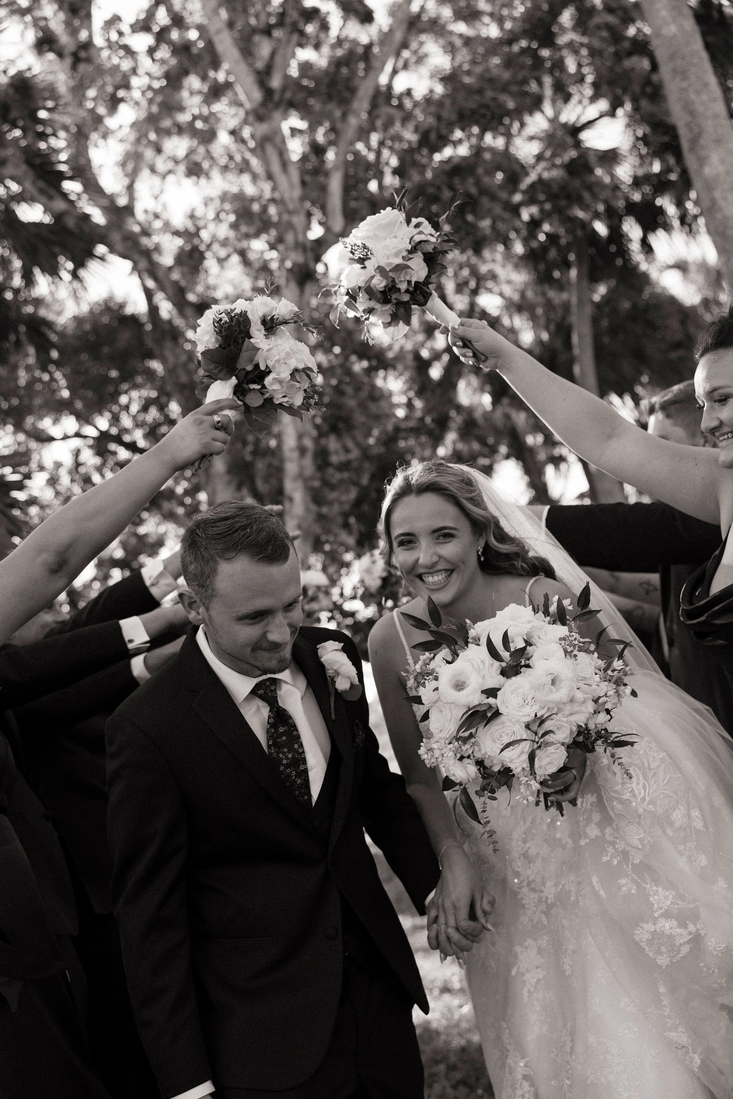 Black and white photo of a bride and groom holding hands, surrounded by bridesmaids and groomsmen holding bouquets, in an outdoor setting with trees.