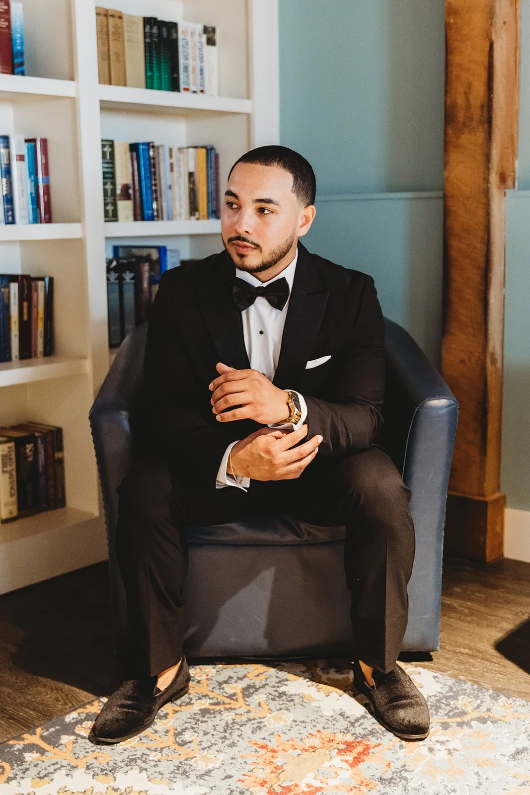A man dressed in a black tuxedo with a white shirt and black bow tie, sitting on a gray armchair in front of a bookshelf filled with books.