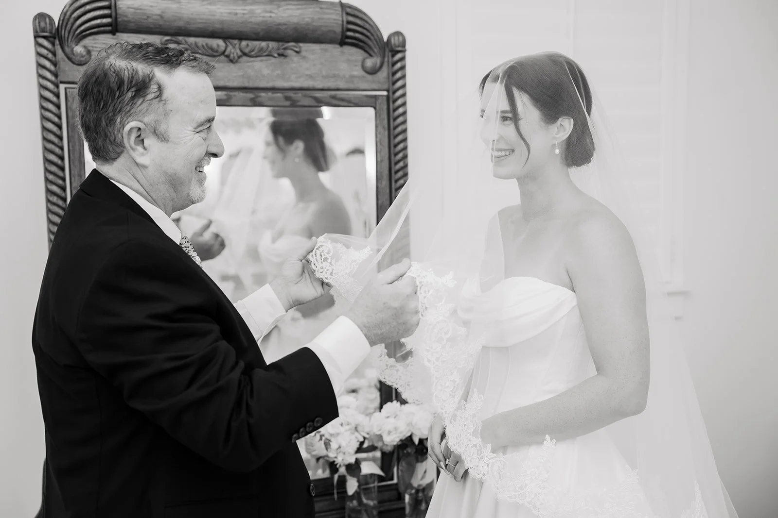 A bride and an older man, likely her father, smile at each other as he helps her with her wedding dress lace. They are indoors, with a mirror and floral arrangement in the background.