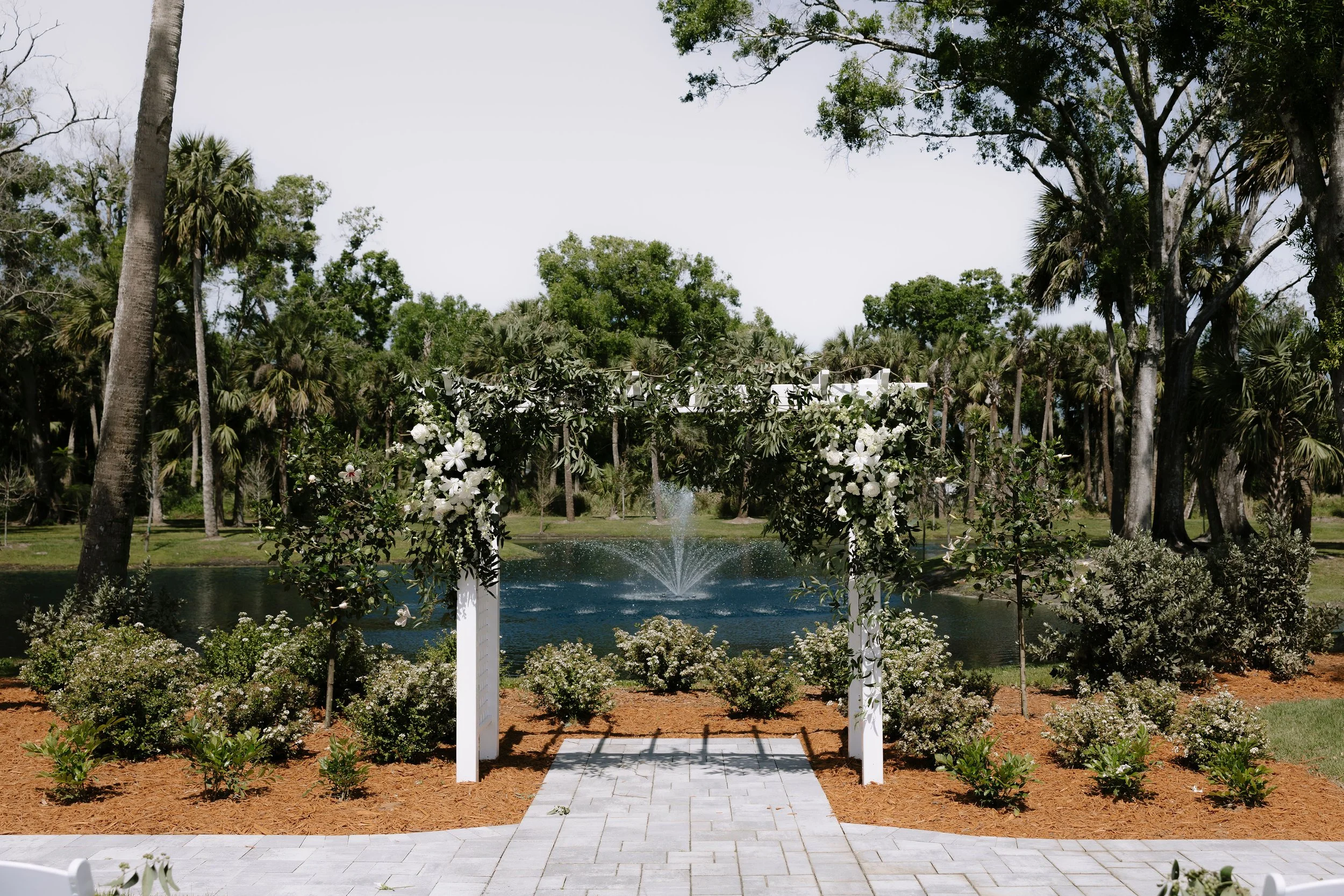 Wedding arbor decorated with white flowers and greenery, with a pond and fountain in the background, surrounded by trees and bushes.