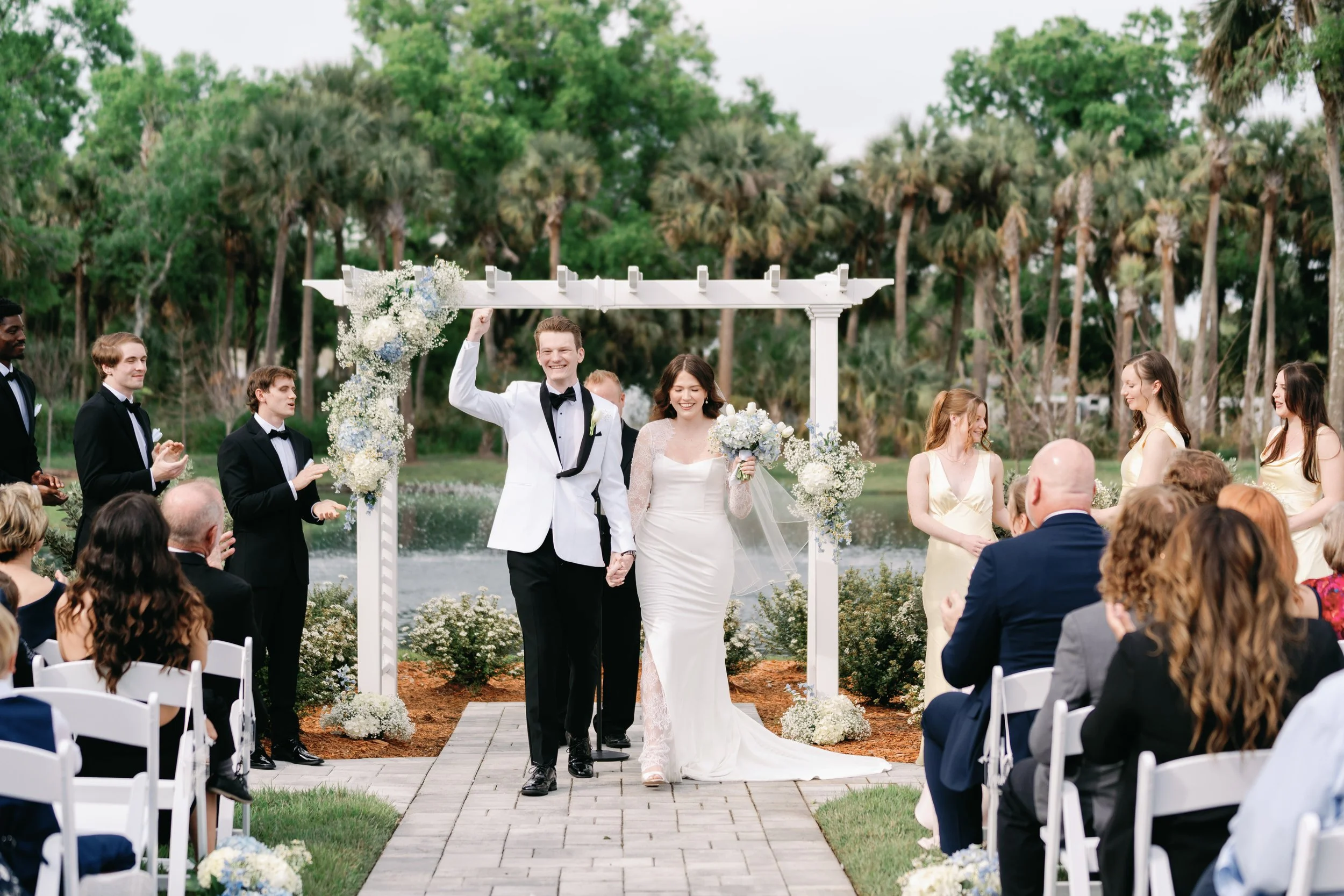 A newlywed couple walks down the aisle at an outdoor wedding ceremony near a body of water, with friends and family clapping and celebrating around them.