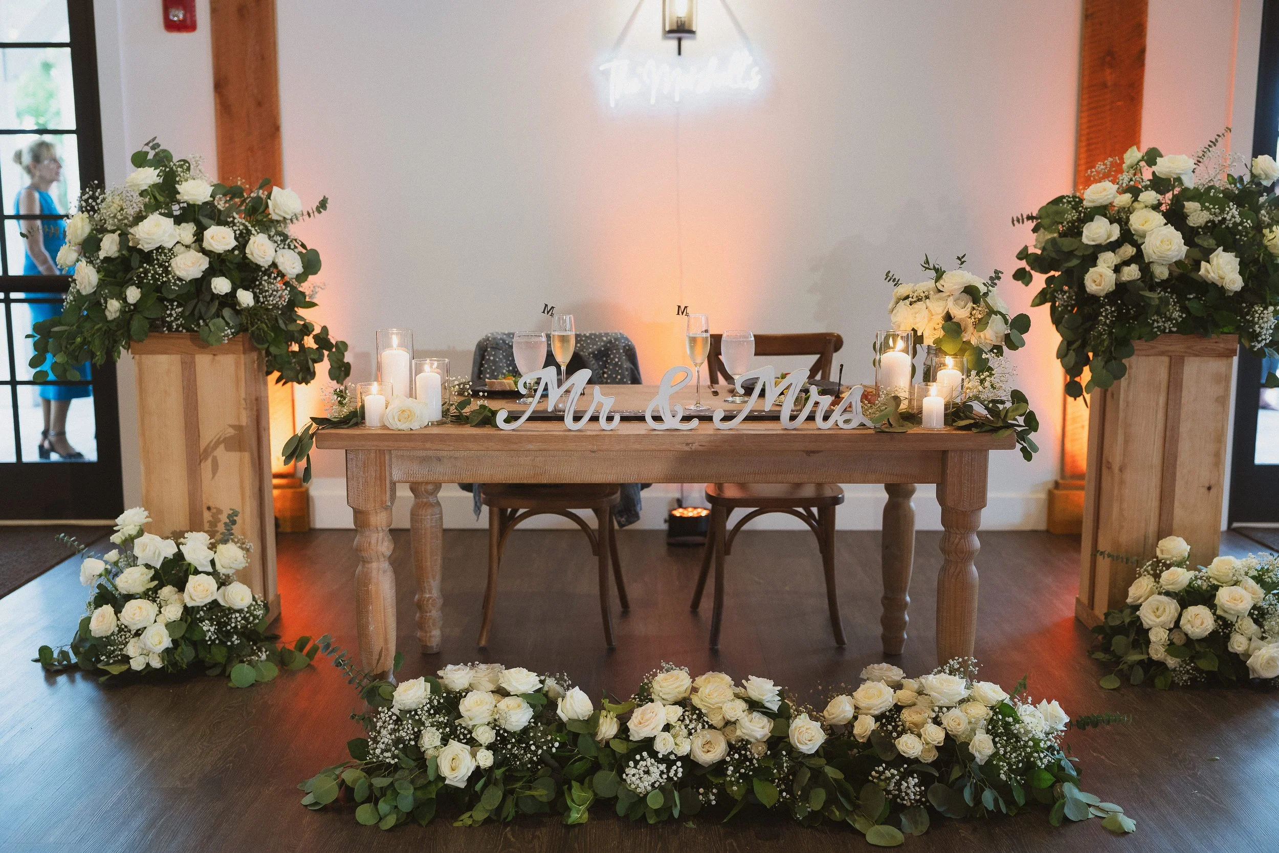 Decorated wedding reception table with floral arrangements, candles, and 'Mr & Mrs' sign, set against a white wall with a neon sign and warm lighting.