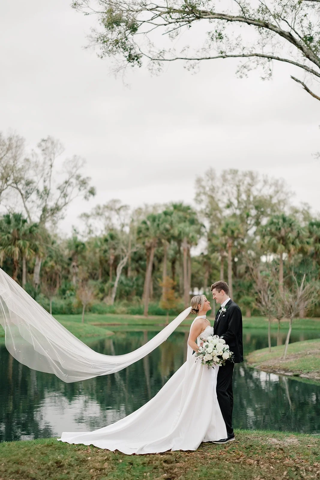 A bride and groom stand by a pond, dressed in wedding attire, with the bride holding a bouquet of white flowers. The bride's long veil flows in the wind. Trees and greenery surround the scene on a cloudy day.