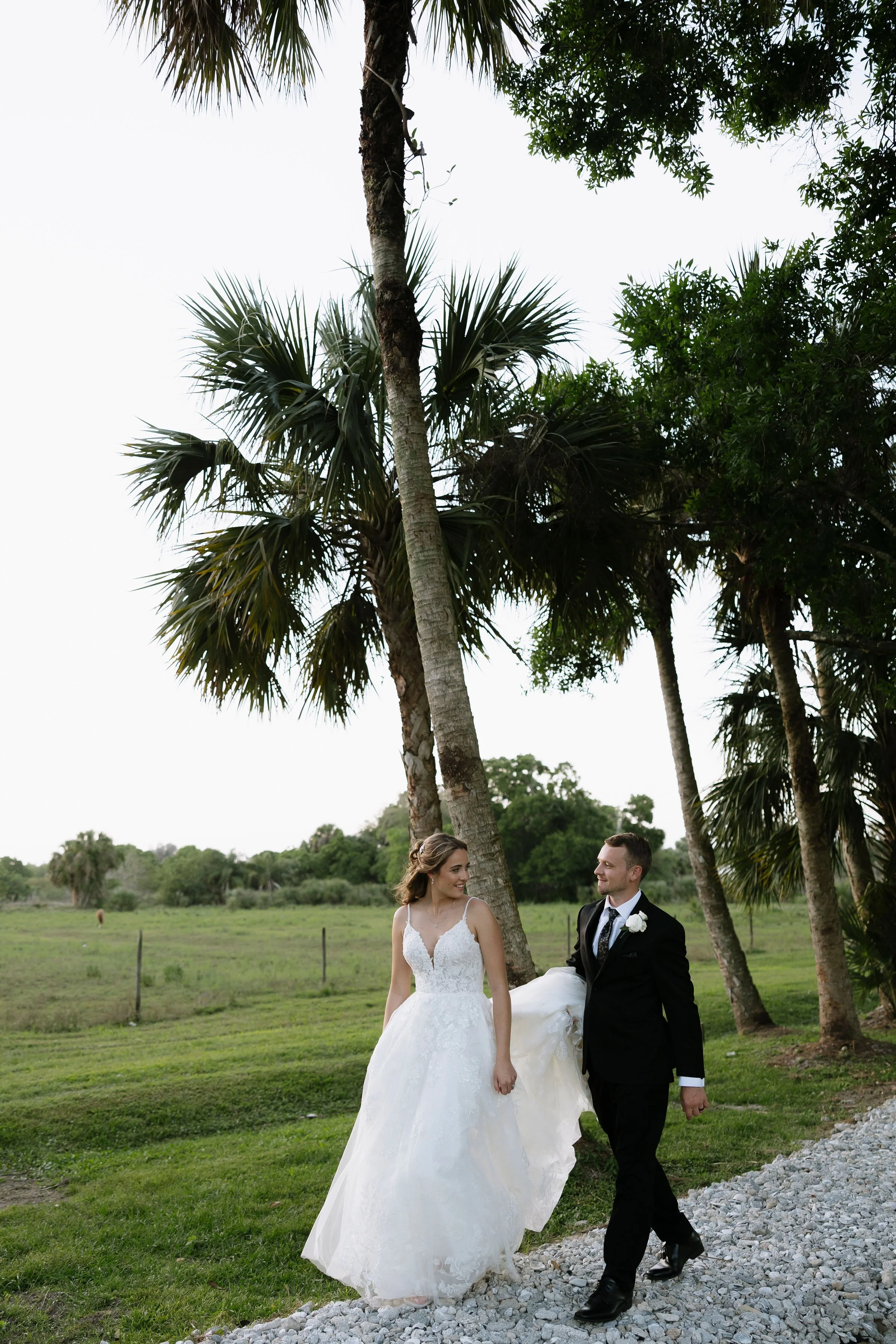 A bride and groom walking outdoors on a grassy area with palm trees, smiling at each other.
