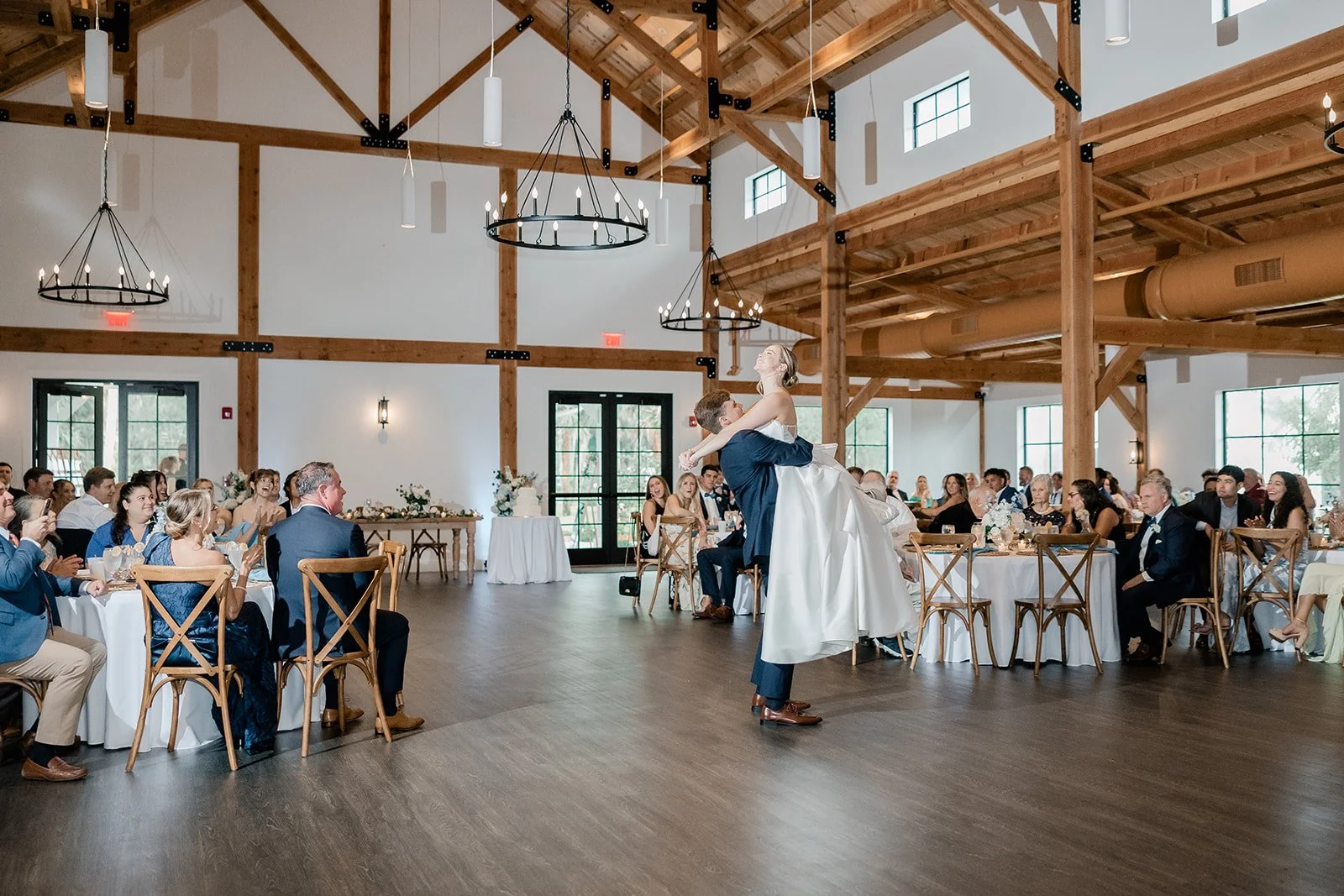 A wedding reception with a bride and groom dancing in the center of the room, surrounded by seated guests inside a rustic hall with wooden beams and large windows.