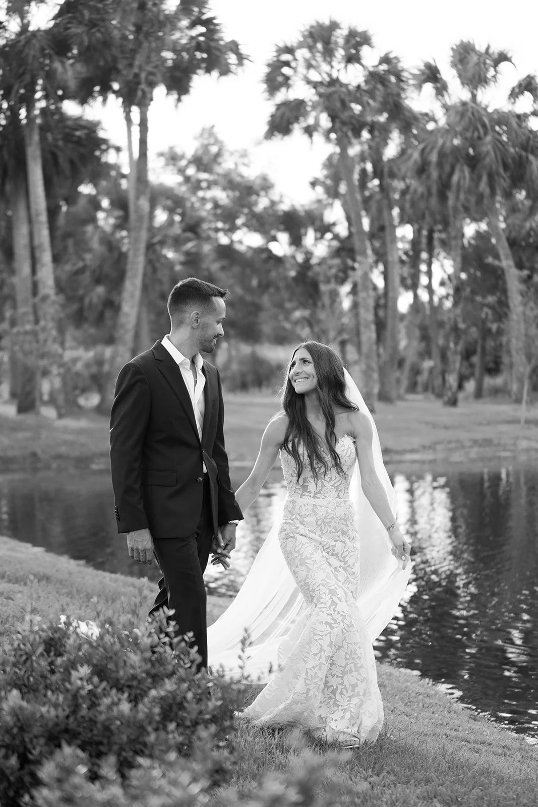 A black and white photo of a bride and groom walking hand in hand near a lake with palm trees in the background, smiling at each other.
