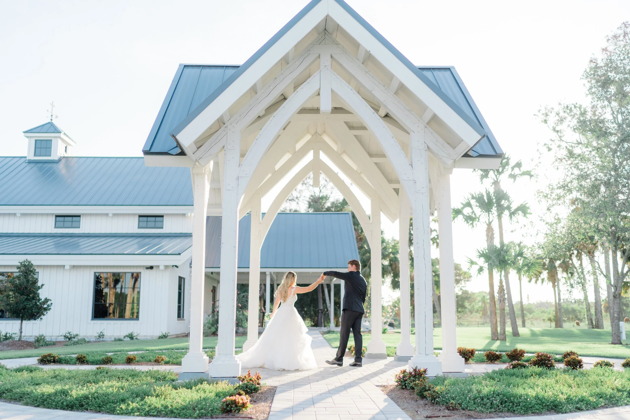 A bride and groom dancing under a white wooden gazebo on a sunny day with a white building and trees in the background.