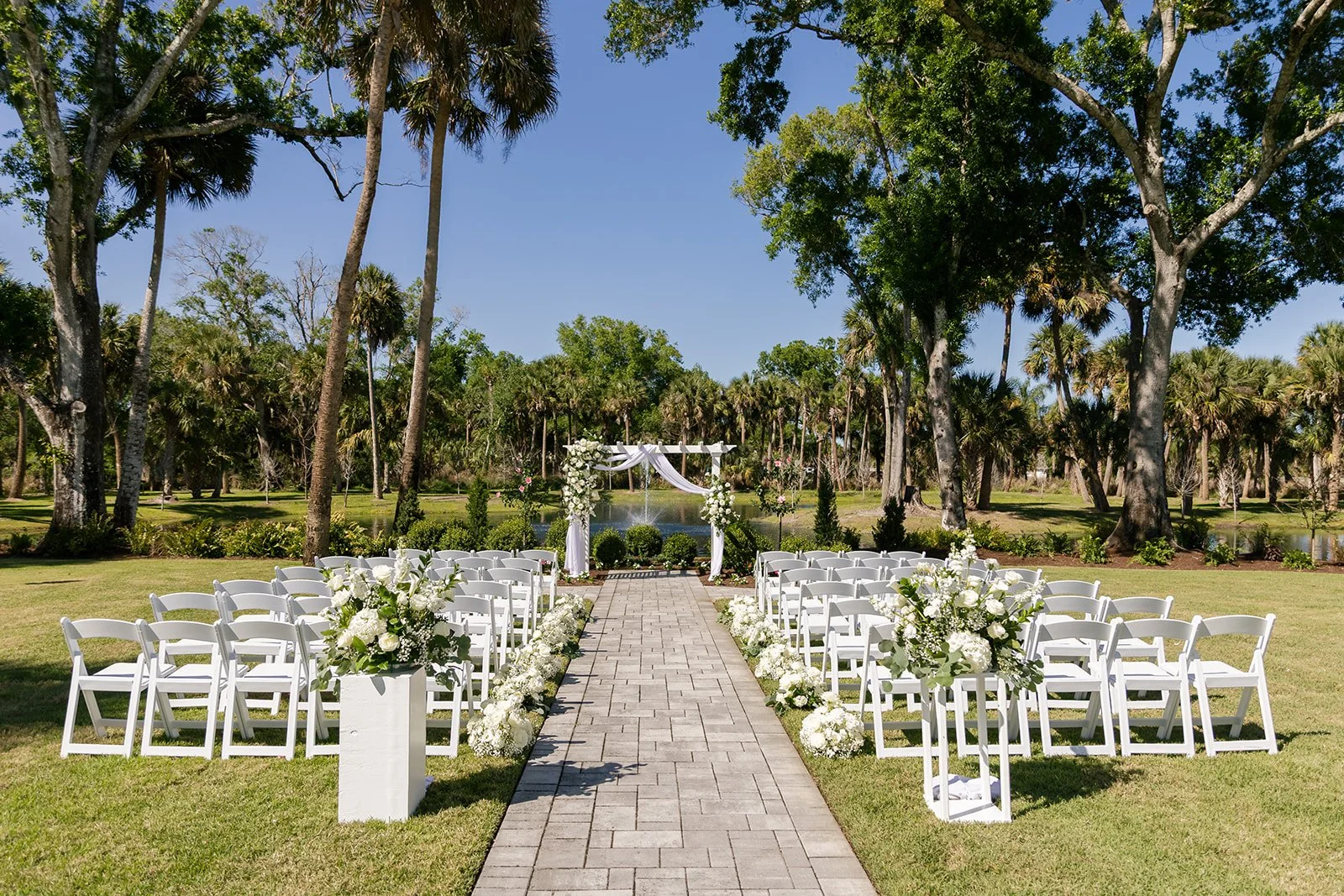 Outdoor wedding ceremony setup at Celebration Grove in Vero Beach with white chairs, floral arrangements, and a decorated arch in a lush green park with tall trees and a pond in the background.