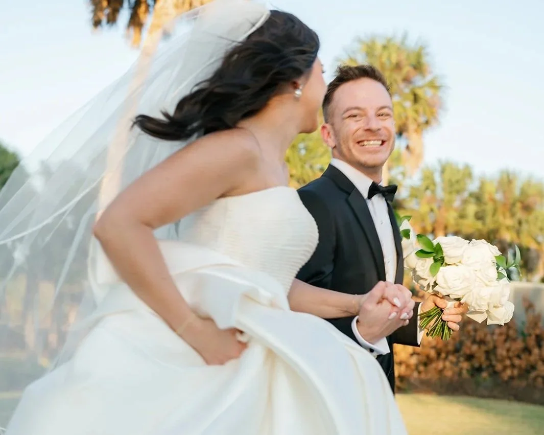 Bride and groom holding hands outdoors on the lush landscaped grounds at Celebration Grove Wedding and Event Venue, smiling, with the bride dressed in a white wedding gown and veil, and the groom in a tuxedo holding a bouquet of white flowers.