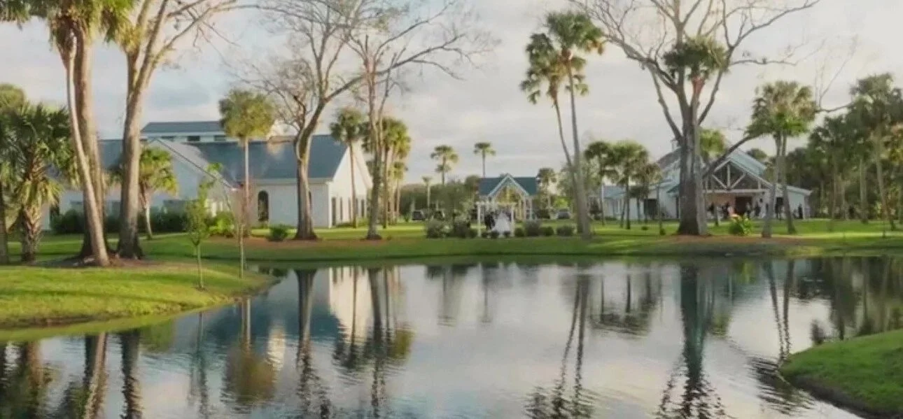 White barn-style building with metal roof, large windows, trimmed in black, surrounded by landscaped garden with trees and bushes
