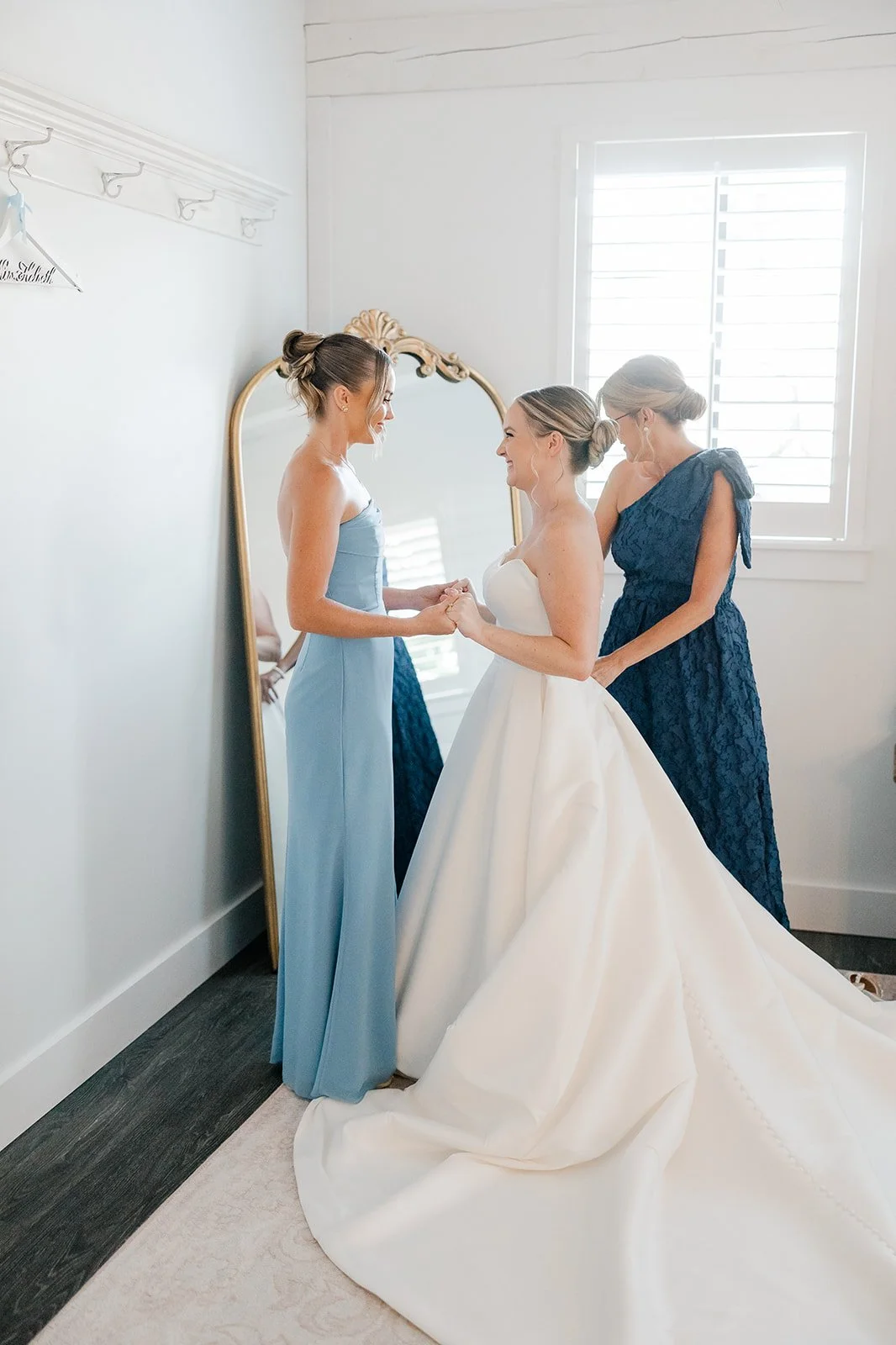 A bride in a white wedding gown holding hands with a bridesmaid in a light blue dress, while another bridesmaid in a dark blue dress stands nearby, in a well-lit room with a mirror and window.