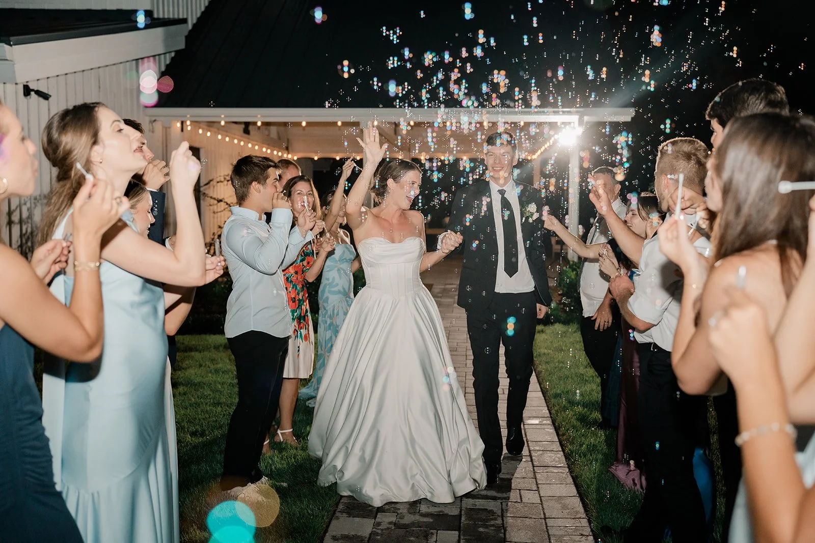 A newlywed couple walking down a lit outdoor path at night, surrounded by friends and family celebrating with bubbles and smiles.