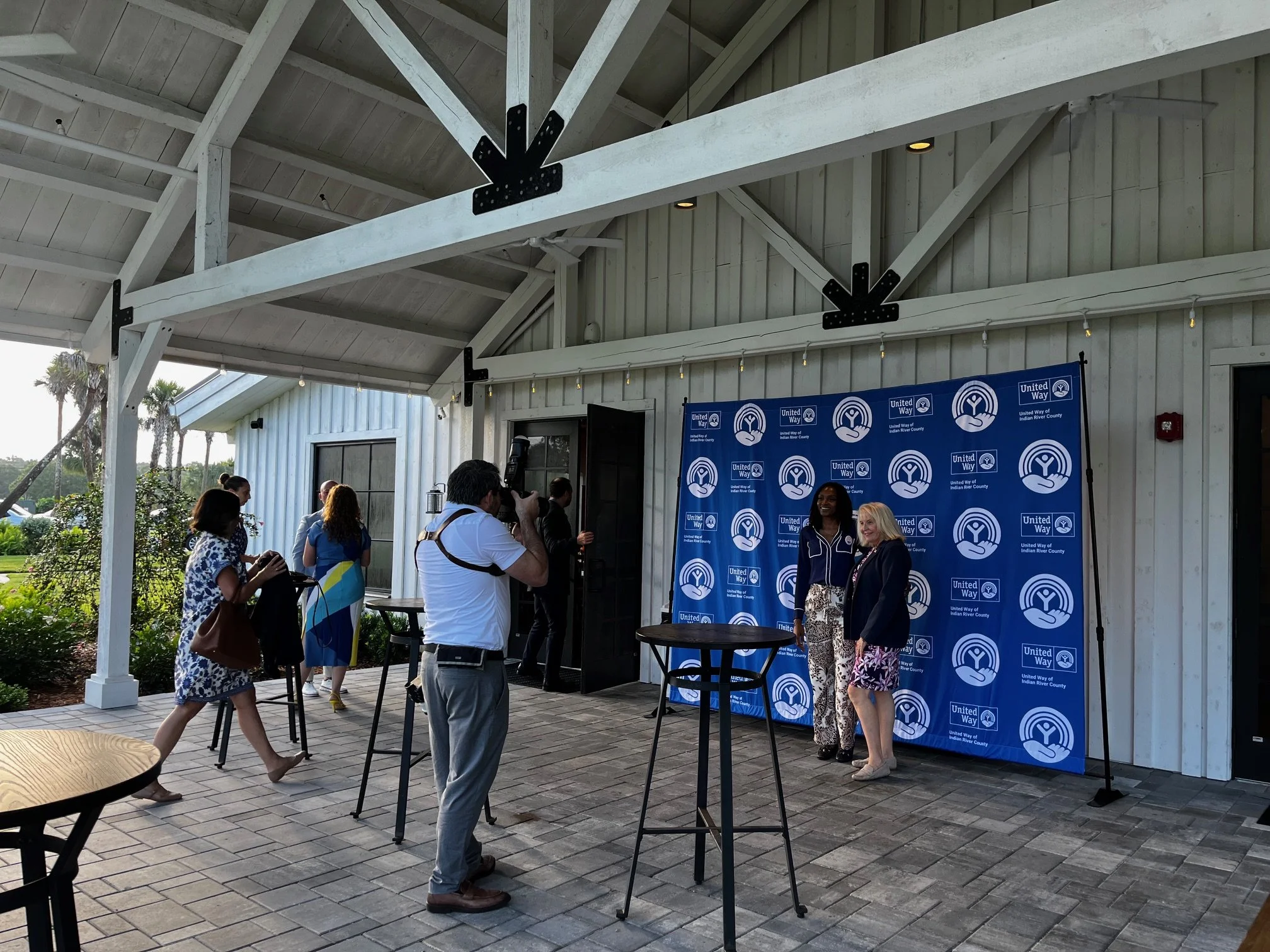 People attending a photo event with a United Way backdrop, some getting their photos taken, others waiting or taking pictures, under a white pavilion with outdoor scenery visible in the background.