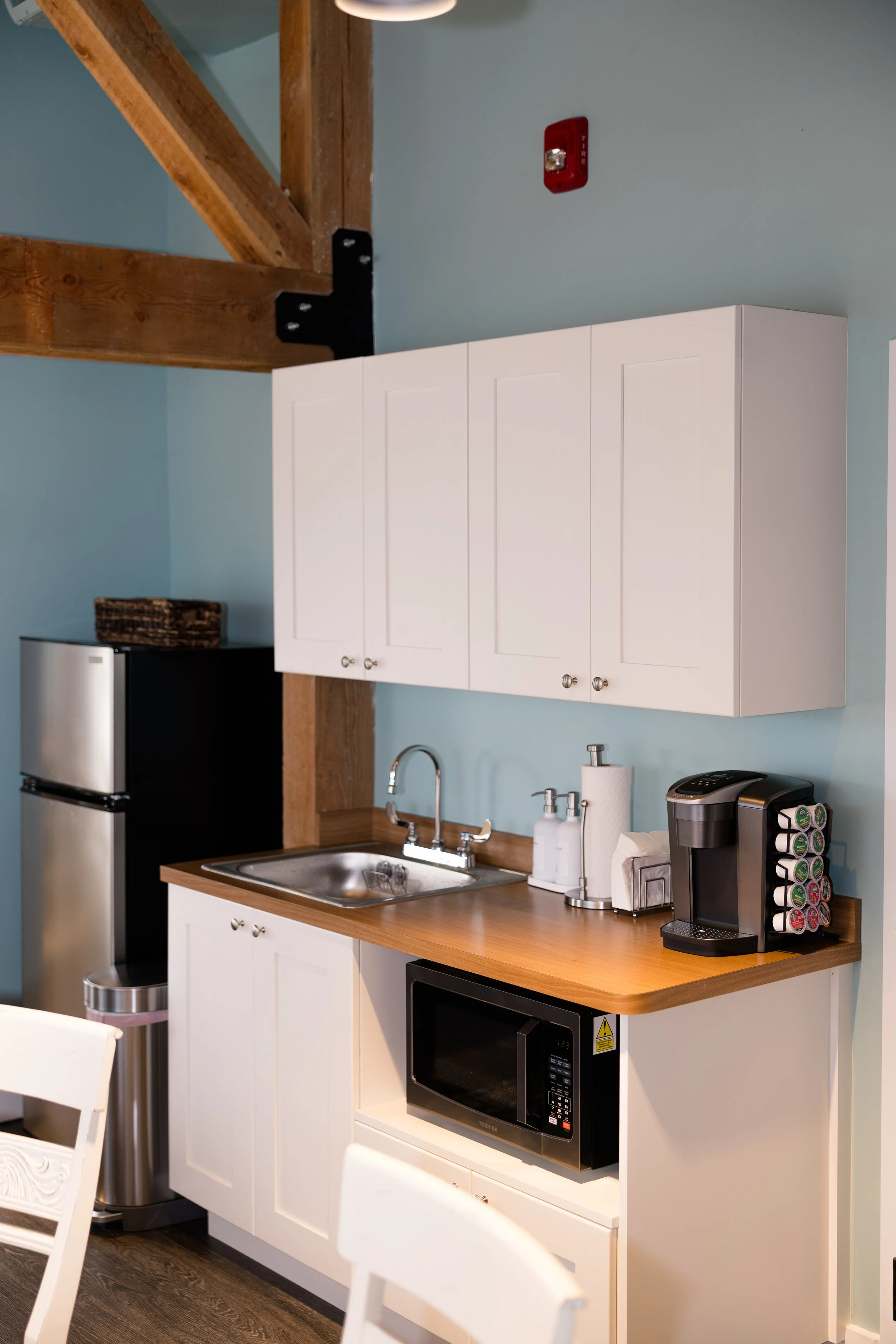 Kitchen area featuring white cabinets, a small sink, a microwave, a Keurig coffee maker, and a refrigerator, with light blue walls and wooden accents.