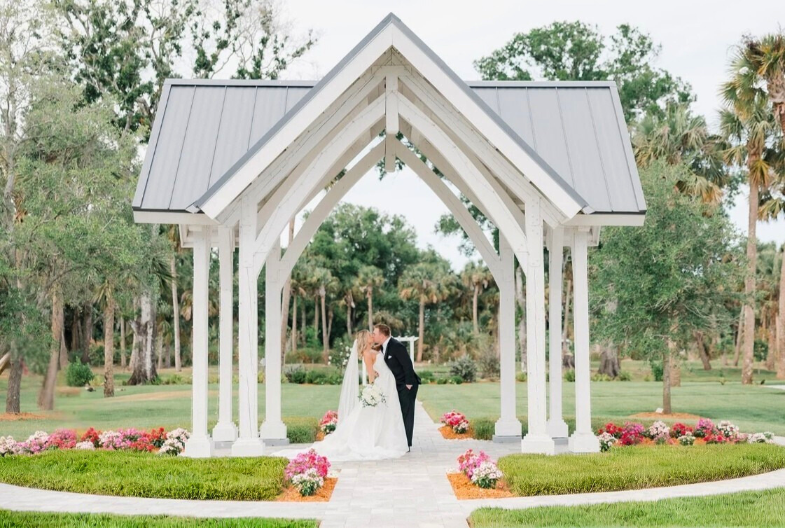 Bride and groom stand beneath the Celebration Grove archway in Vero Beach, Florida — surrounded by golden light and oak trees, capturing the beauty and sacred joy of their wedding day.