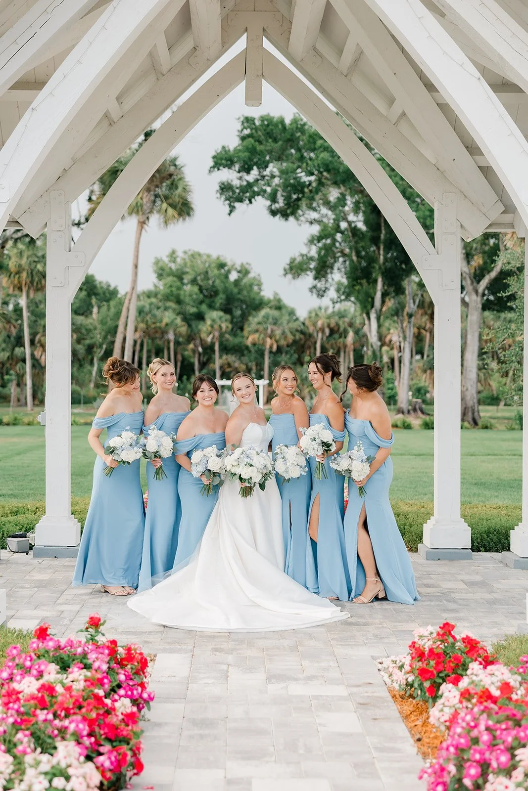 A group of seven women, including a bride in a white wedding dress, standing under a white wooden archway outdoors. The women are wearing matching light blue bridesmaid dresses and holding white bouquets, with a lush green background and colorful flo