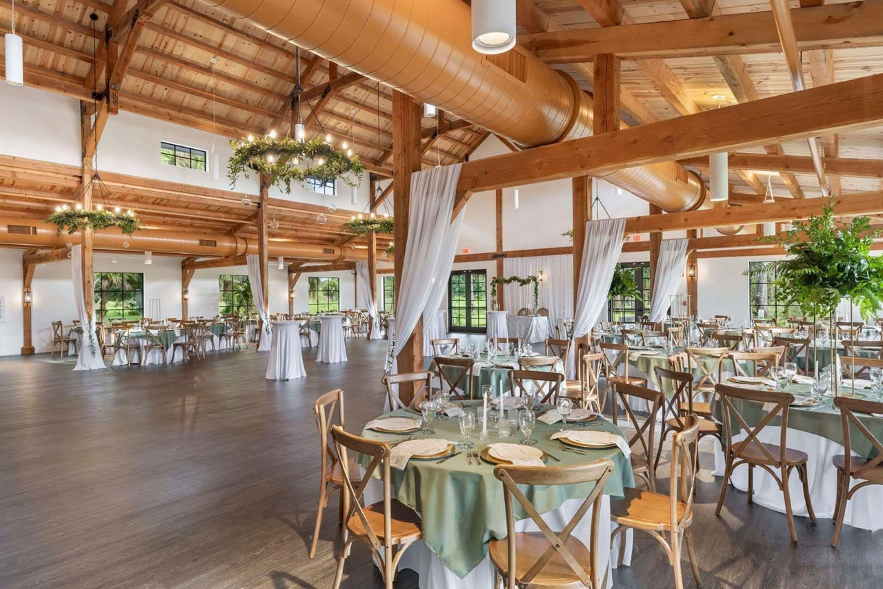 Elegant reception hall with wooden beams, white curtains, greenery decor, and round tables set for a formal event at Celebration Grove in Vero Beach, FL.