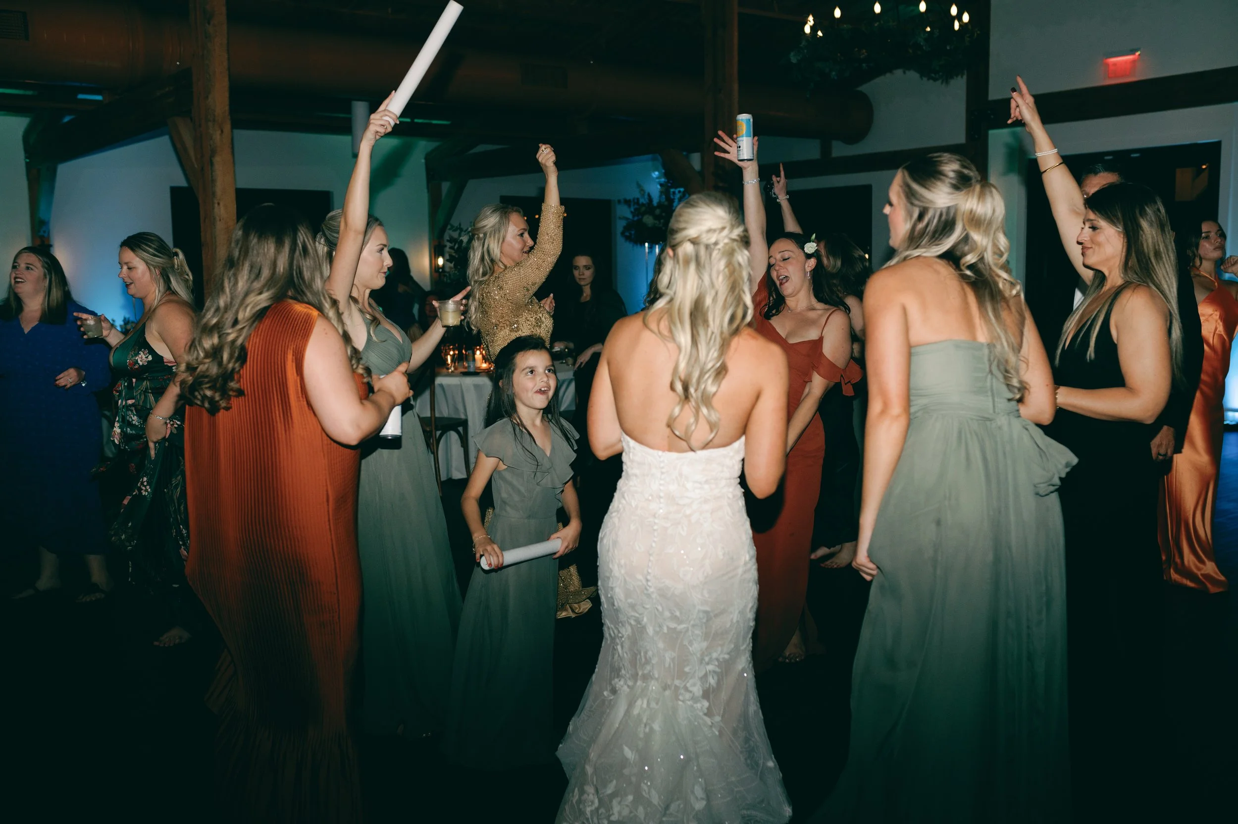 Group of women and girls dancing and celebrating at a wedding reception, with some holding drinks or objects in their hands.