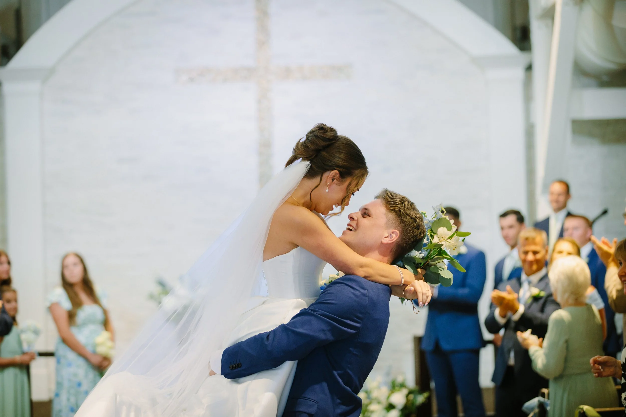 A bride and groom share a joyful moment at their wedding ceremony, with the bride being lifted by the groom, holding a bouquet of flowers, and smiling at each other in front of guests in a bright, elegant venue.