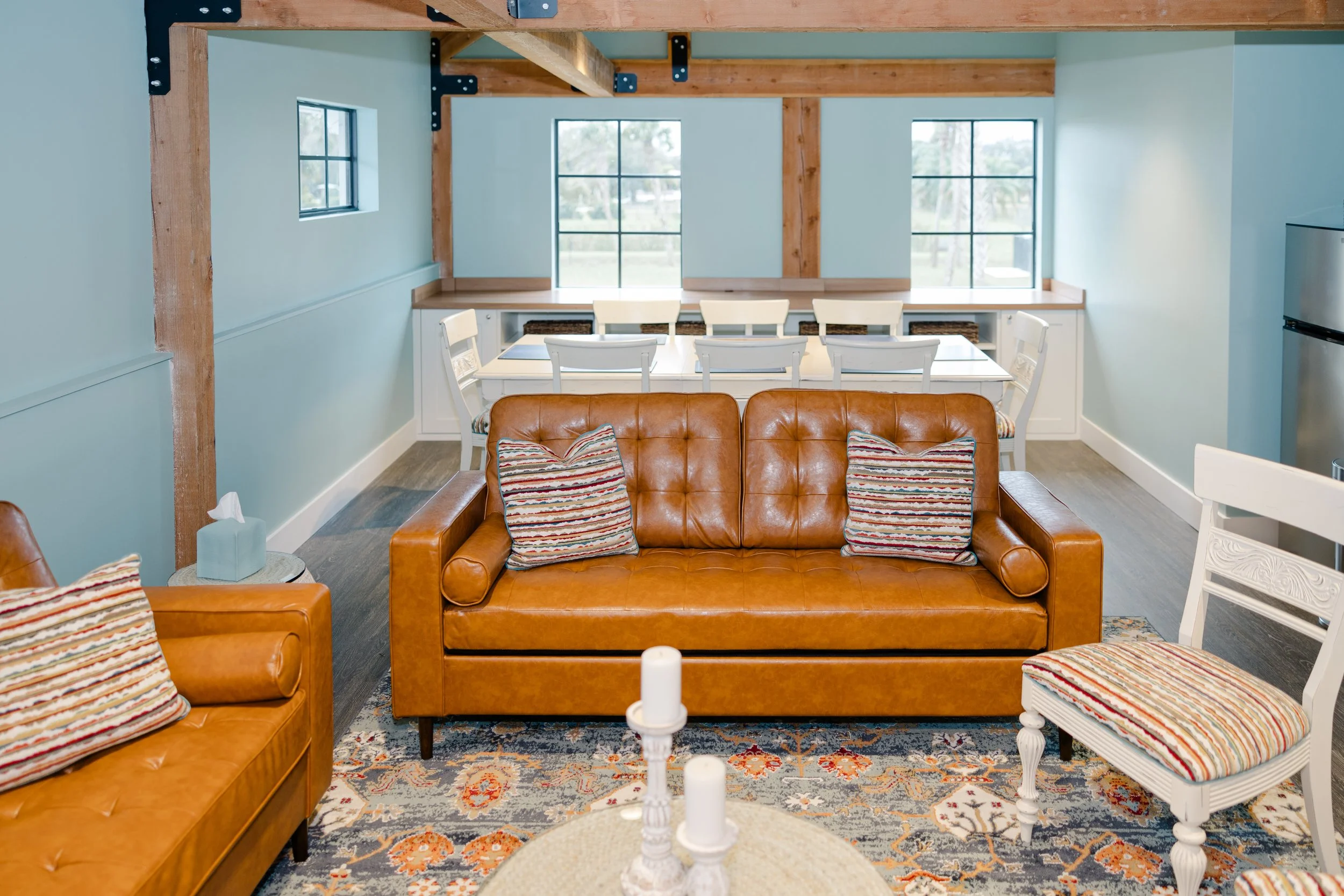 Living room with leather sofas, striped pillows, a white dining table with chairs, and large windows letting in natural light, featuring exposed wooden beams.