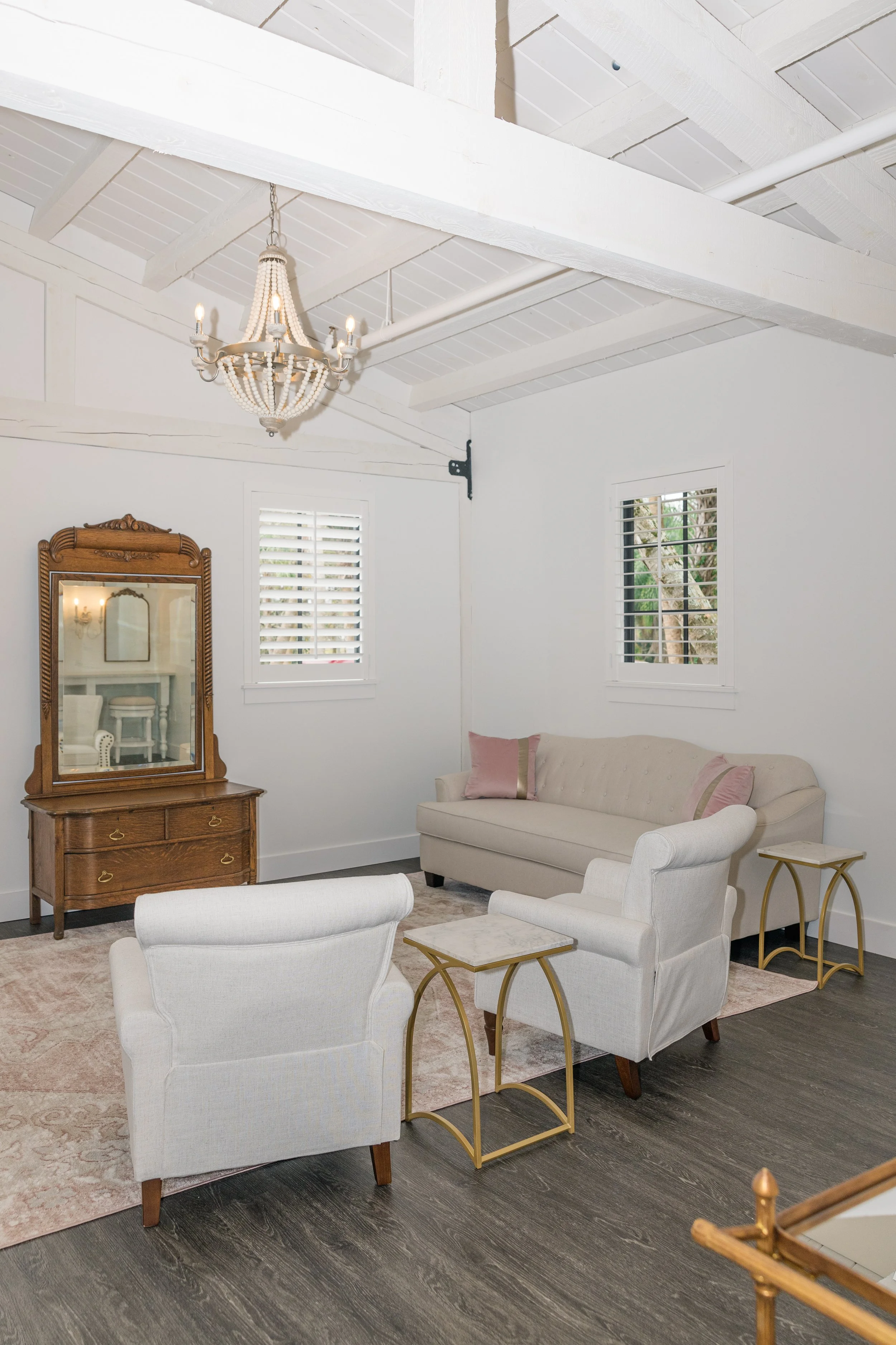 A cozy living room with white walls and ceiling beams, a chandelier, two white armchairs, a beige sofa with pink pillows, gold side tables, a wooden dresser with mirror, and windows with white shutters.