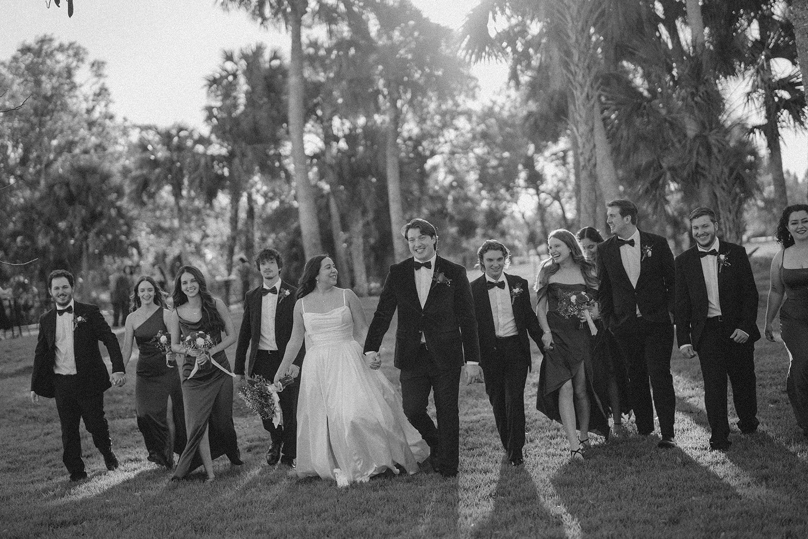 Black and white photo of a wedding party walking hand in hand outdoors in a park with trees. The bride and groom are in the center, smiling, with the bride in a white gown and the groom in a tuxedo. The wedding party includes men and women dressed in