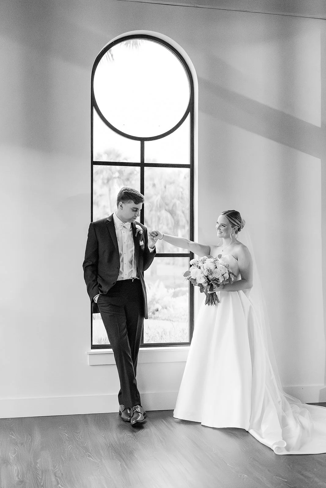 A bride and groom inside a building with large arched window, the bride's arm extended towards the groom, who is looking down at her hand, with the bride smiling. The bride holds a bouquet of flowers, and both are dressed formally.