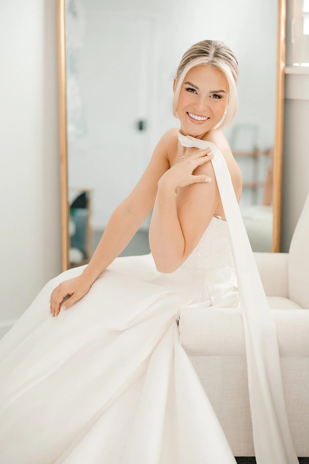 A woman in a white wedding dress smiling and sitting on a beige couch, touching her shoulder with her right hand.