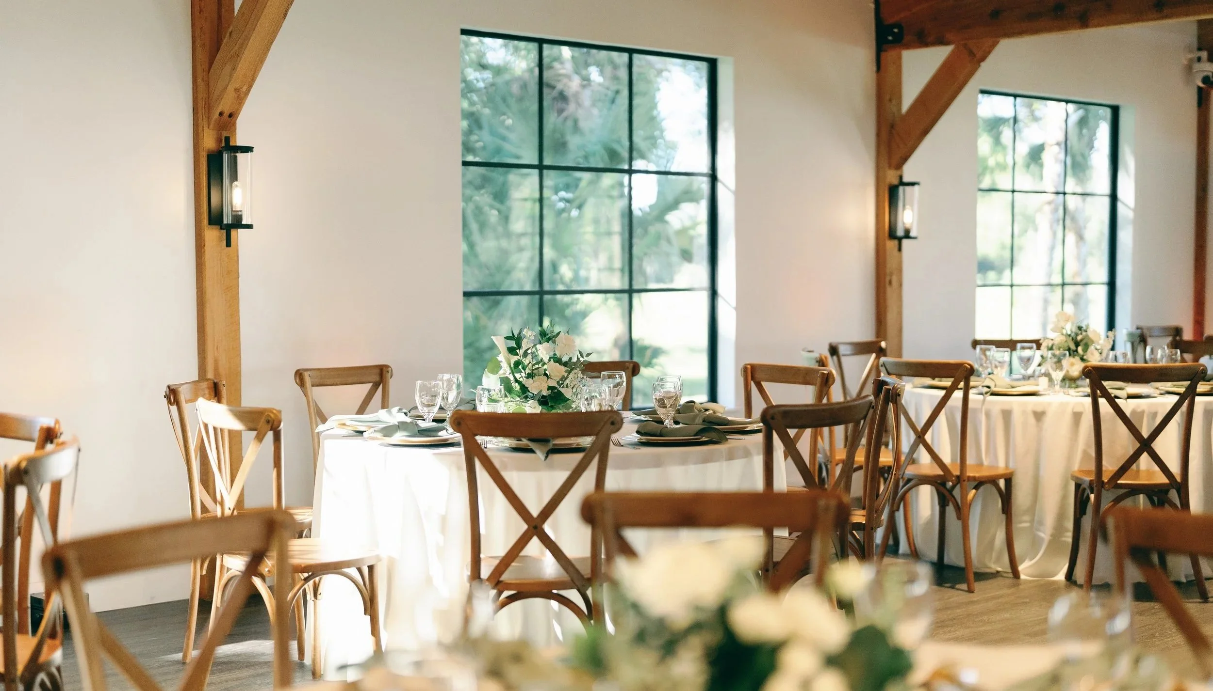 Elegant event space inside the reception hall at Celebration Grove displaying round tables covered in white tablecloths. Each table has floral centerpieces and place settings. Wooden chairs surround the tables. The room is lit by natural light.
