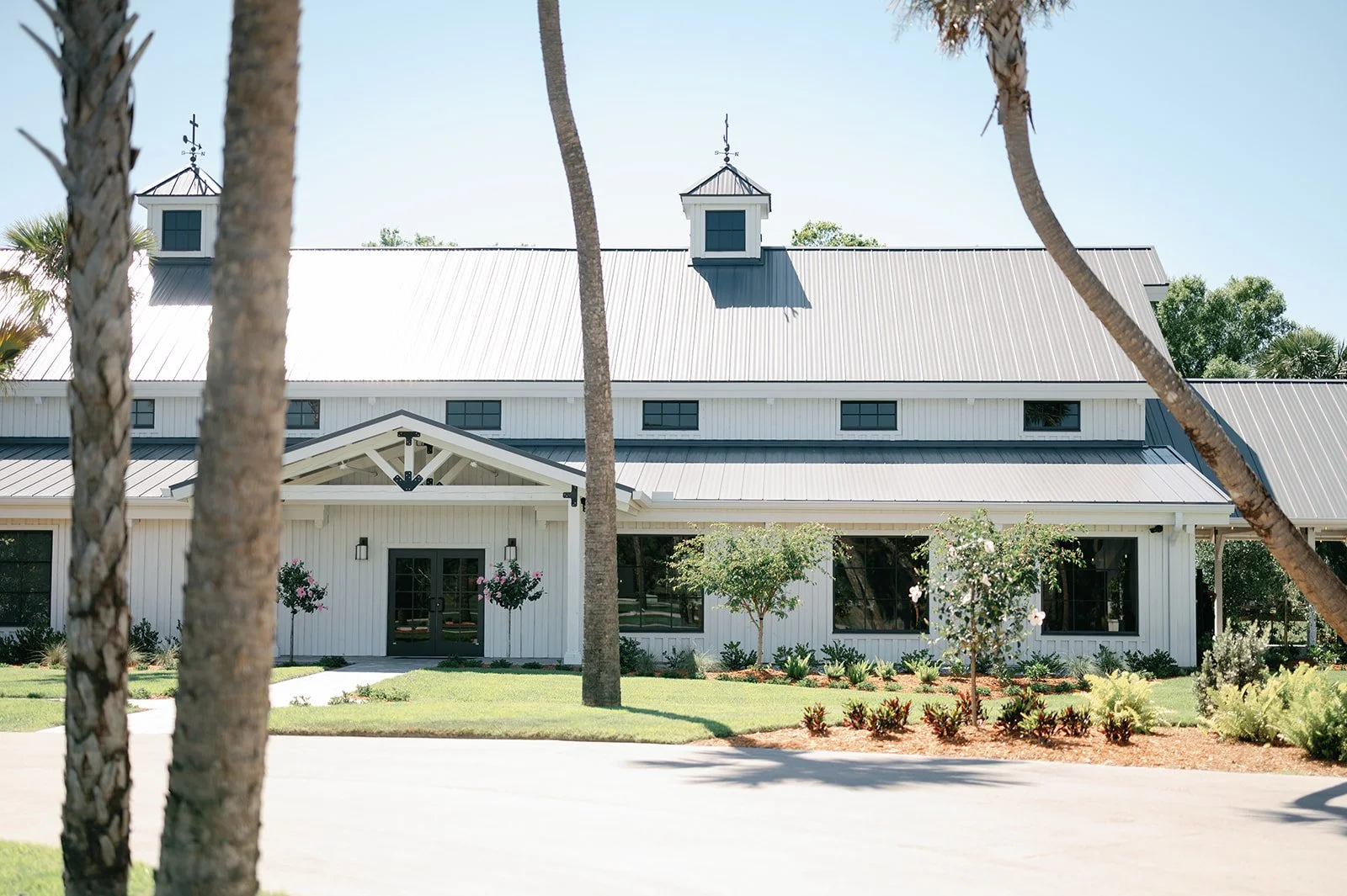 Large white barn-style building with metal roof, surrounded by landscaped garden with trees and bushes, under clear blue sky.