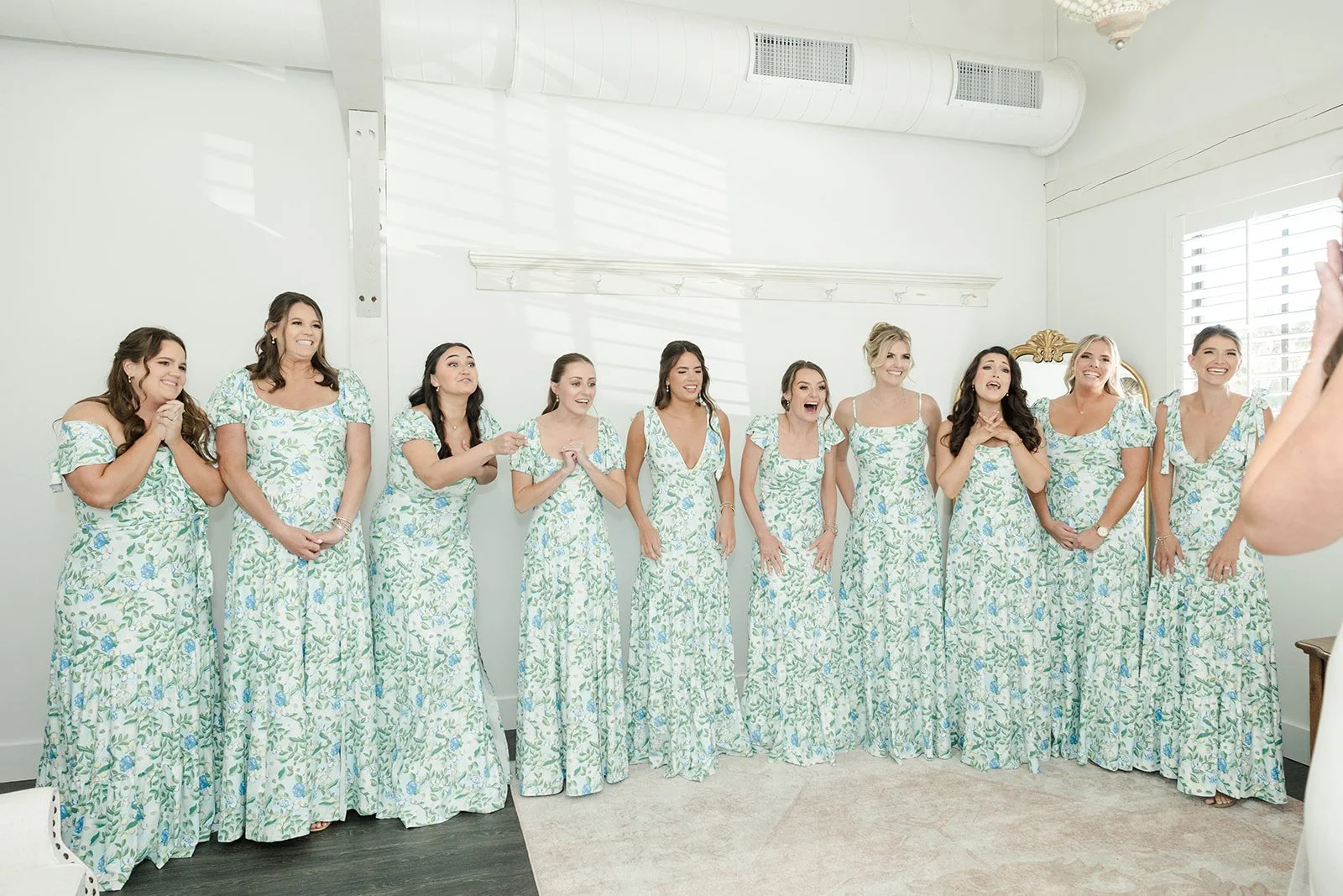 A group of eleven women dressed in matching floral dresses standing indoors, smiling and reacting to someone taking a photo.
