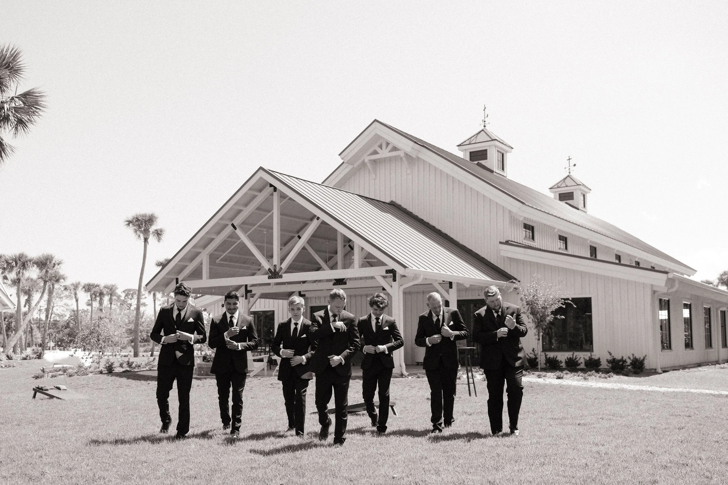 Group of seven men in suits walking on grass outside a large white building with a metal roof, in a sunny outdoor setting with trees.