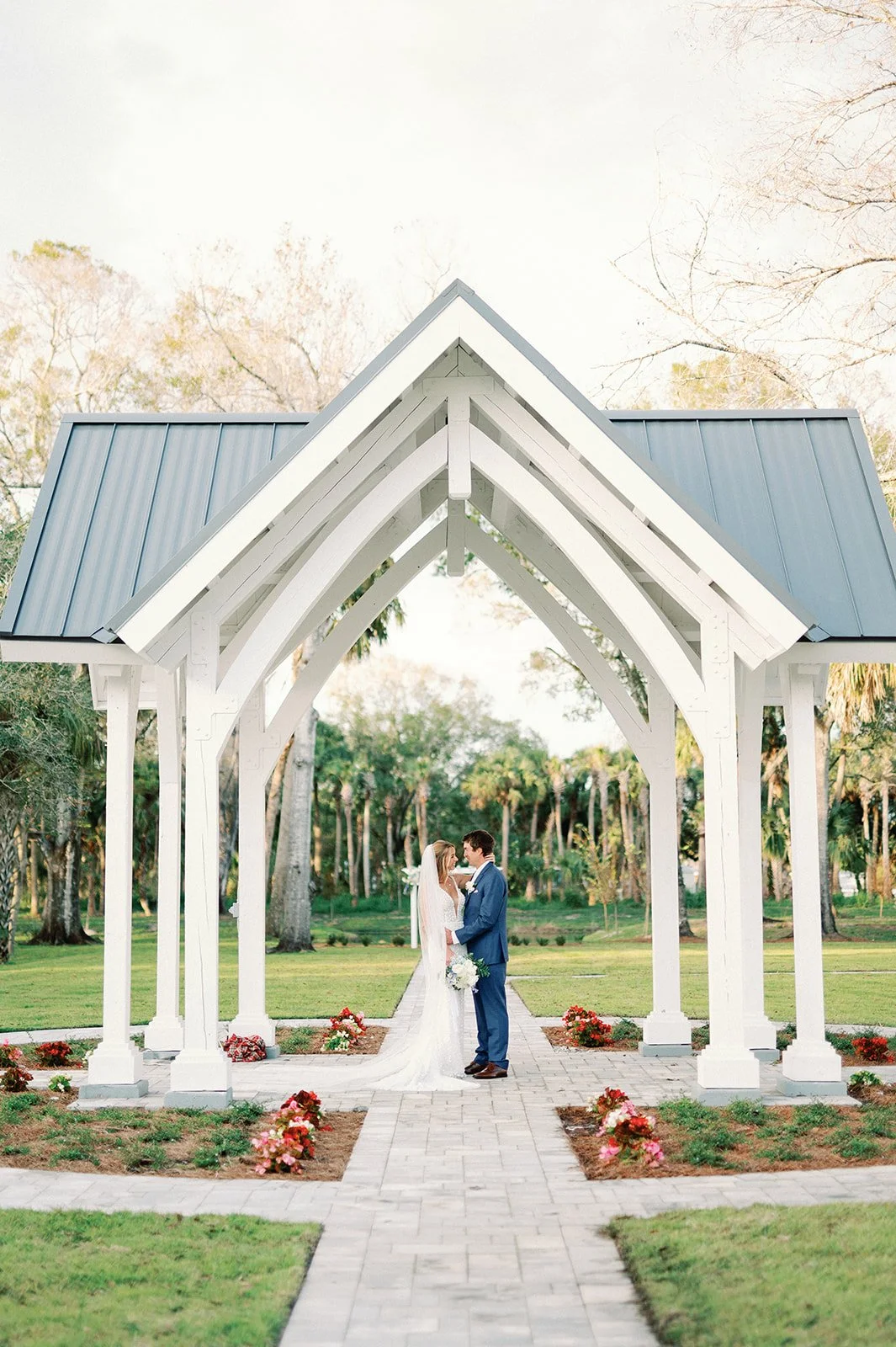 A bride and groom standing under a white wooden archway with a gray metal roof, in a park with trees and greenery in the background, during a wedding ceremony.