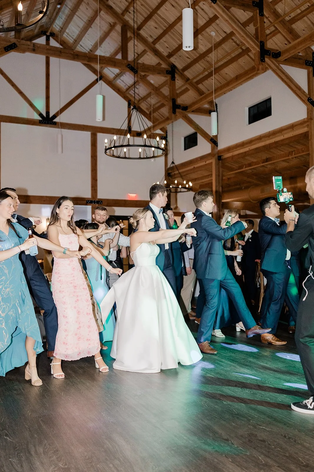 People dancing at a wedding reception inside a barn, with a wooden ceiling and dim lighting.
