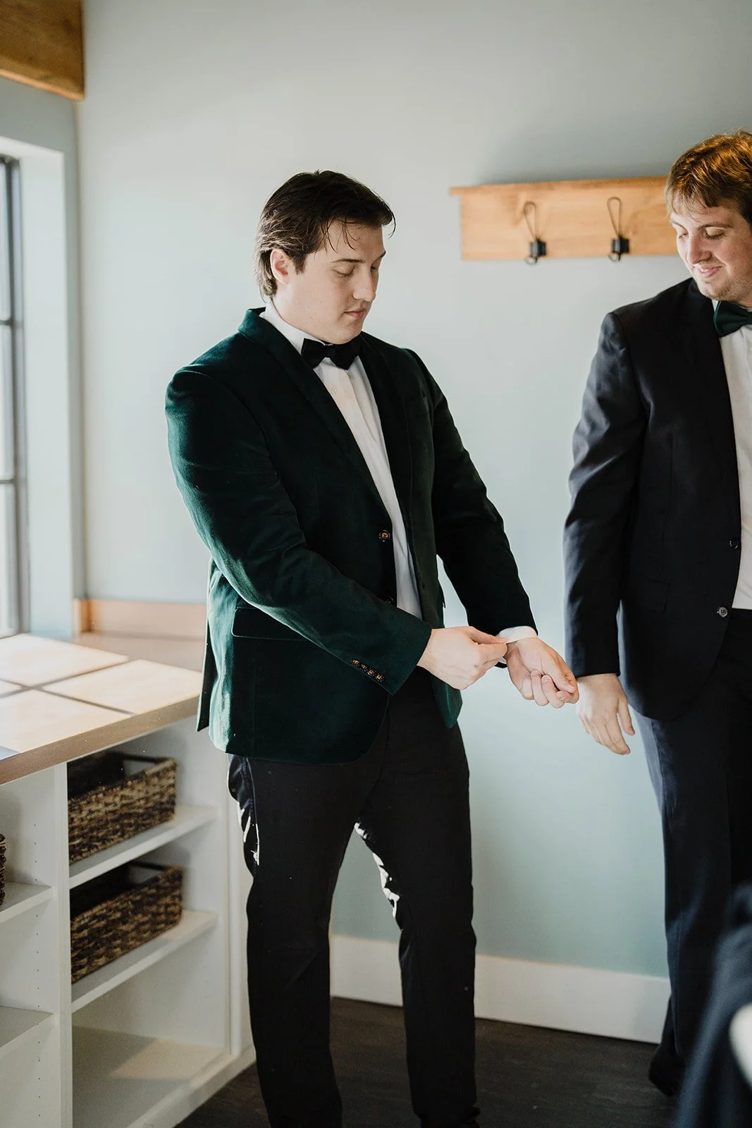A man in a black tuxedo putting on a wristwatch, standing next to another man in a tuxedo, in a room with sunlight coming through a window.