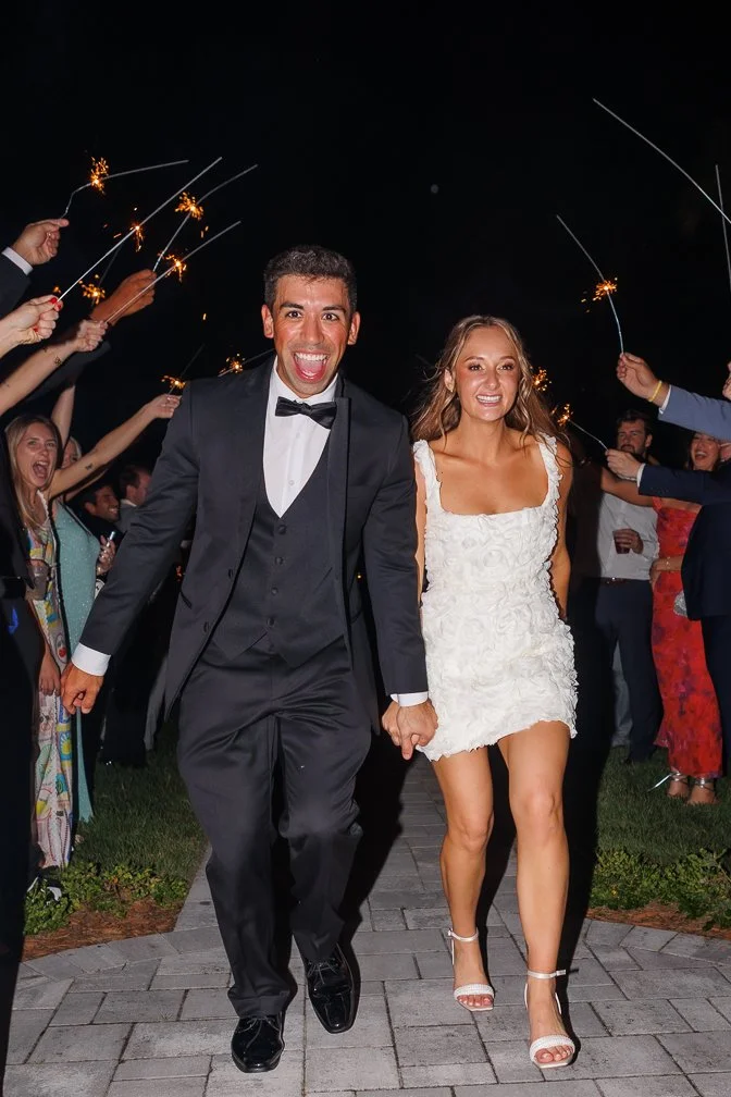 A newlywed couple, a groom in a tuxedo and a bride in a white dress, holding hands and smiling as they walk together at night during a celebration with sparklers. Guests are surrounding them, cheering and waving sparklers.