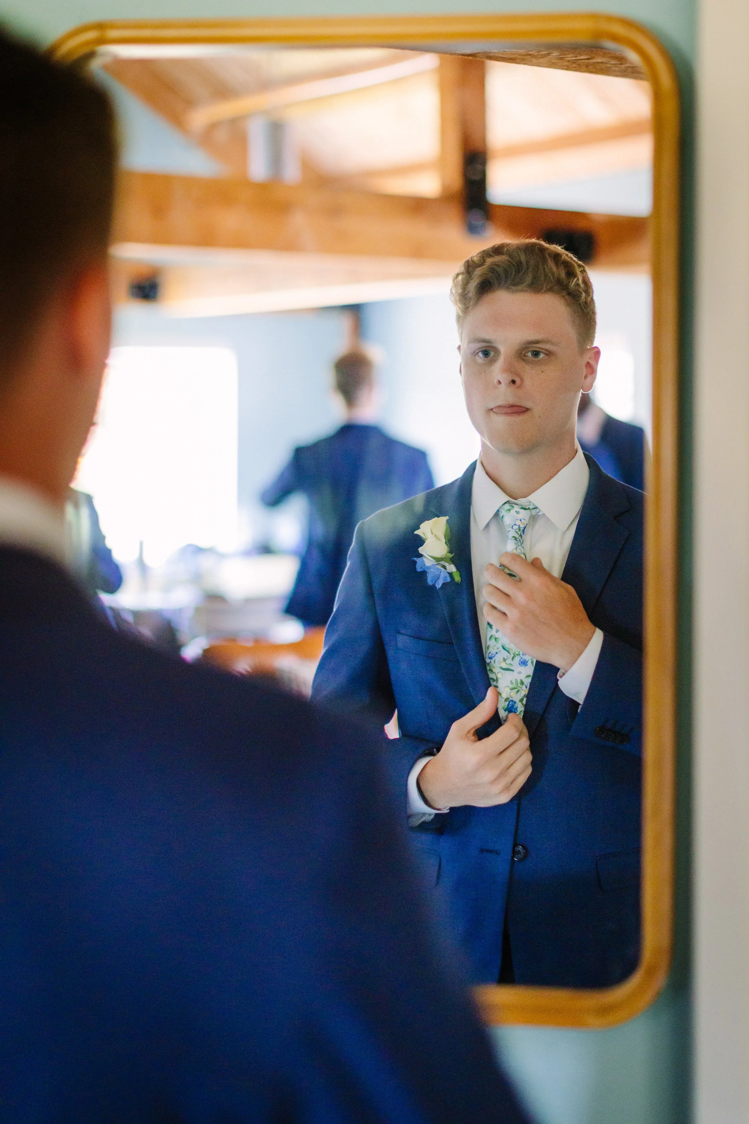 A young man in a blue suit and floral tie looking at himself in a mirror, adjusting his jacket, with a boutonniere on his lapel, preparing for a formal event or wedding.