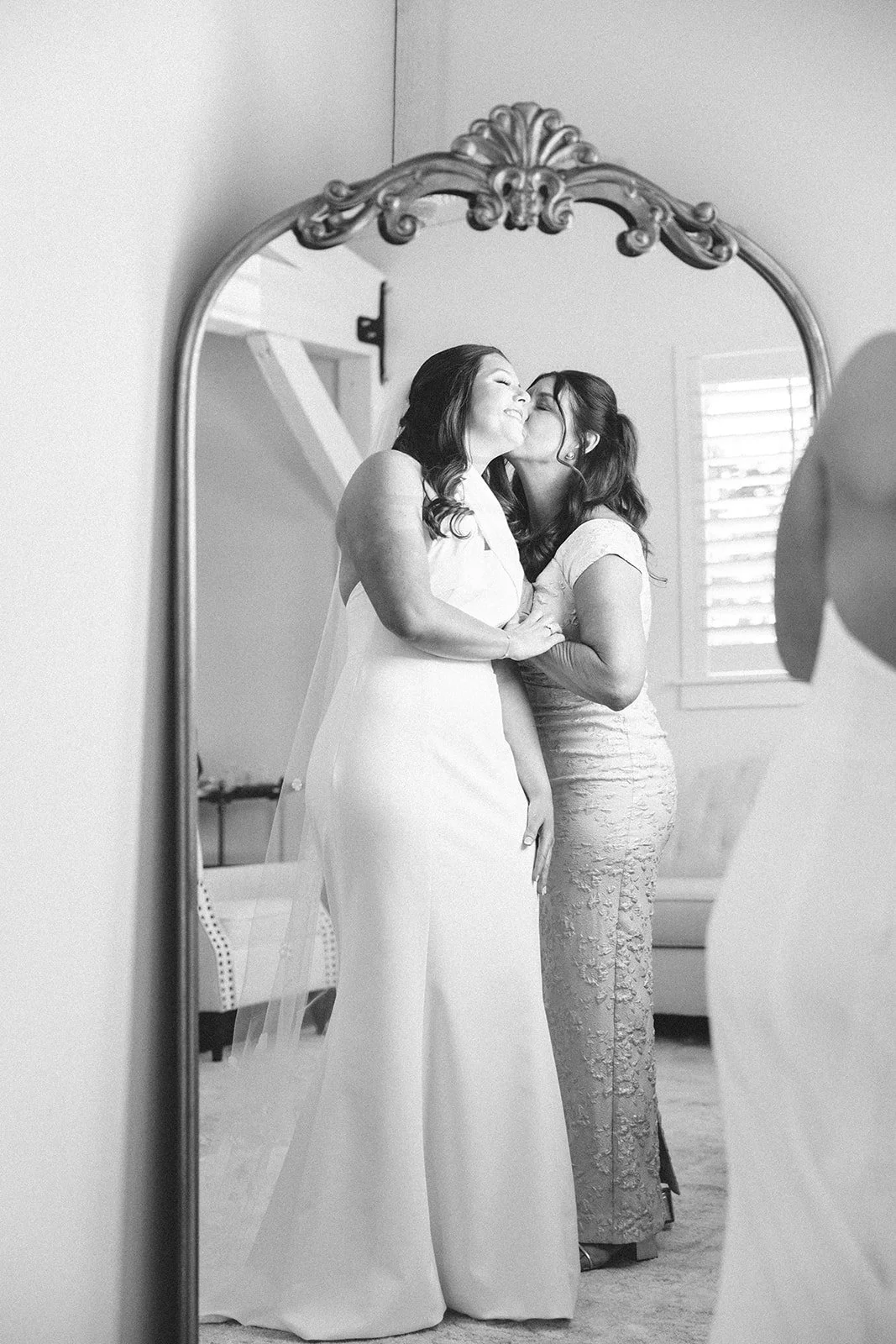 Two women share a tender moment in front of a mirror, one in a wedding dress and the other in a lace gown, with a window with shutters in the background.