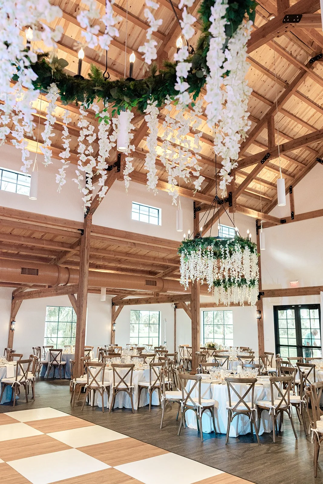 A spacious wedding reception hall decorated with hanging white floral arrangements and green foliage chandeliers. The room features high wooden ceilings, large windows, and round tables covered with white tablecloths, set for dining.