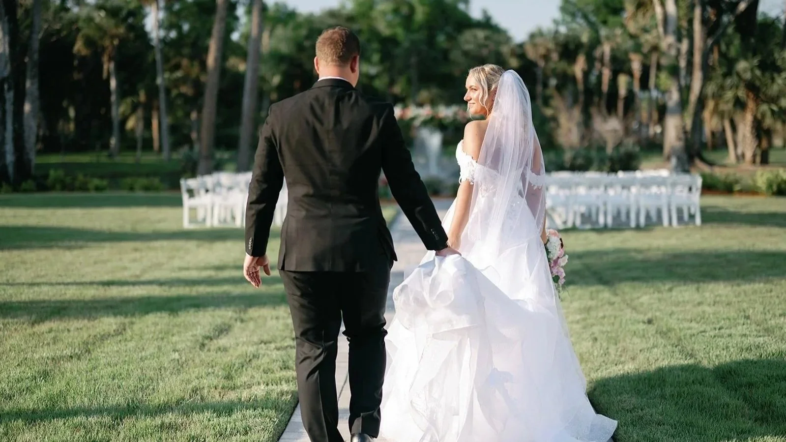 A bride in a white wedding dress and veil holding a bouquet and holding hands with a groom in a black suit walking across a grassy lawn at an outdoor wedding ceremony setup with white chairs and trees in the background.