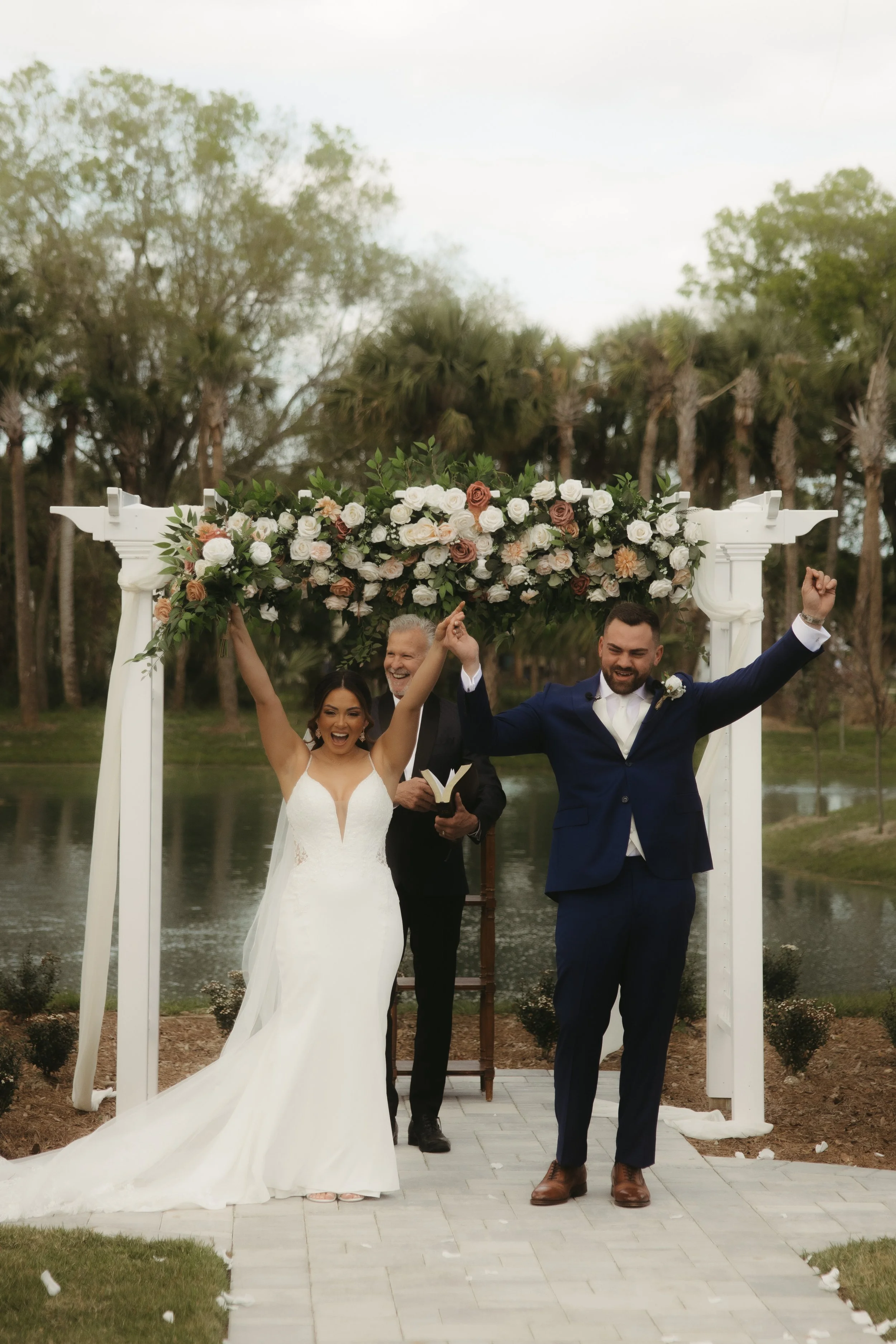 A bride and groom celebrating at their outdoor wedding ceremony under a floral arch, with an officiant behind them, on a paved path by a pond and trees.