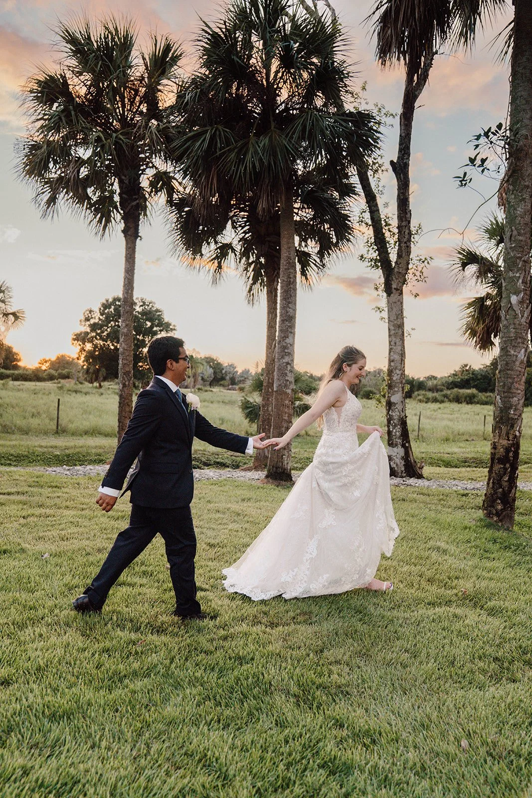 A bride in a white wedding dress playfully pulls her dress as a groom in a dark suit walks towards her outside on a grassy field with palm trees during sunset.