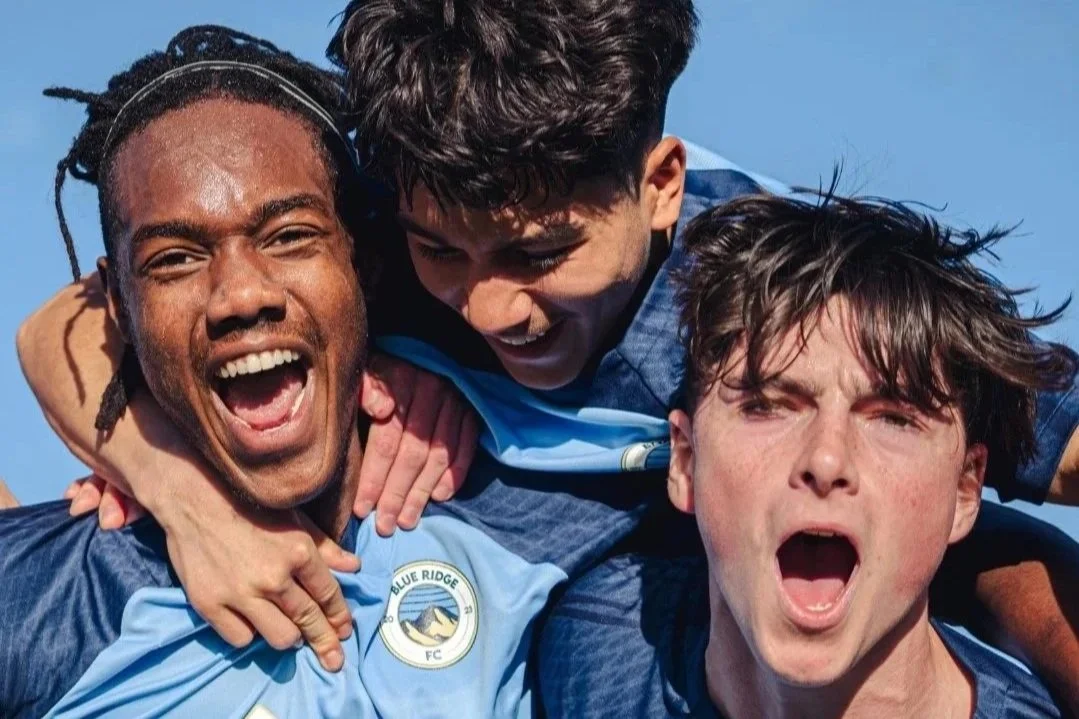 Three young male soccer players in blue jerseys celebrating together with excitement and joy, showing camaraderie and victory.